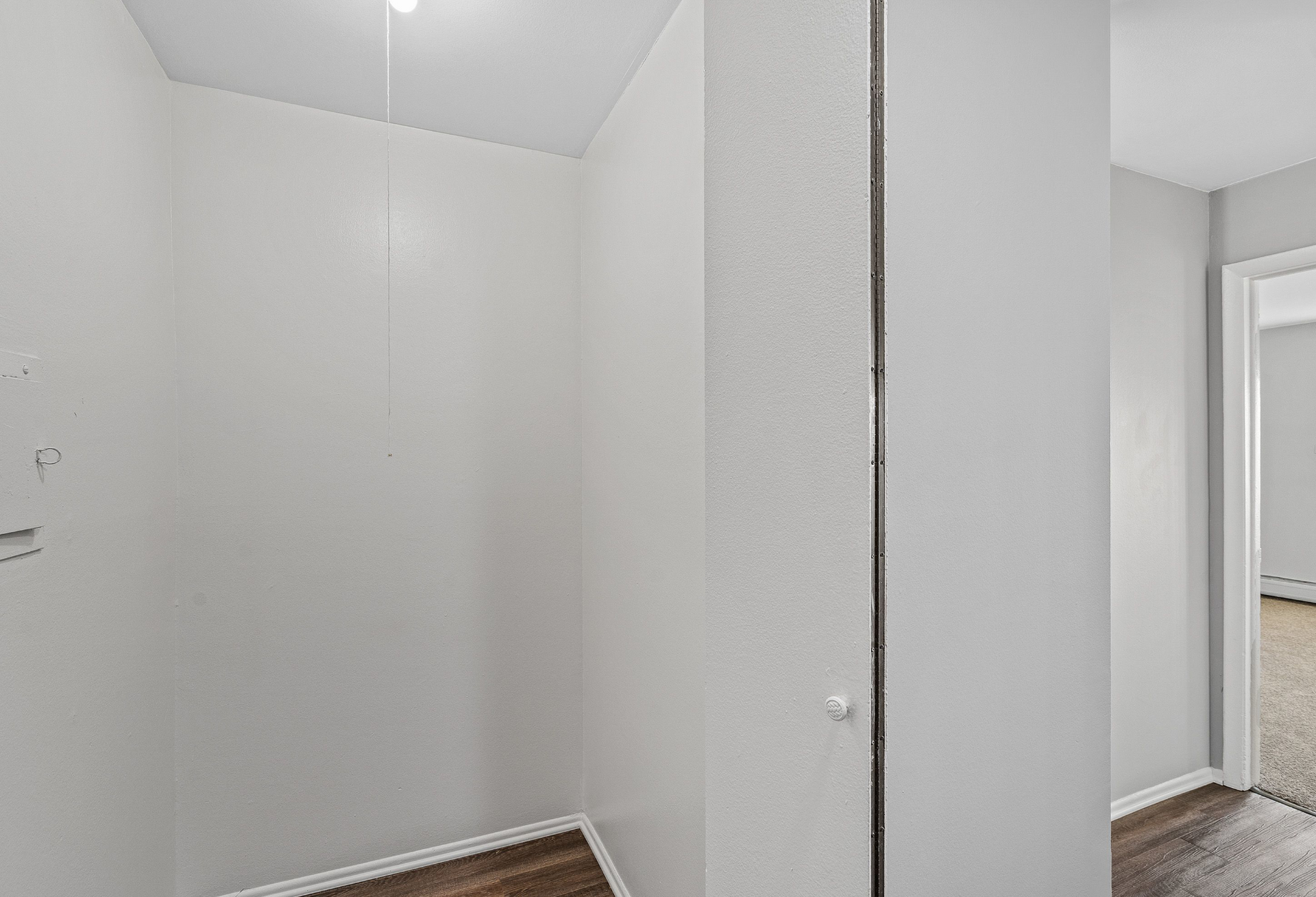 Empty white closet nook with pull-chain light and wood floor, opening to a carpeted room.
