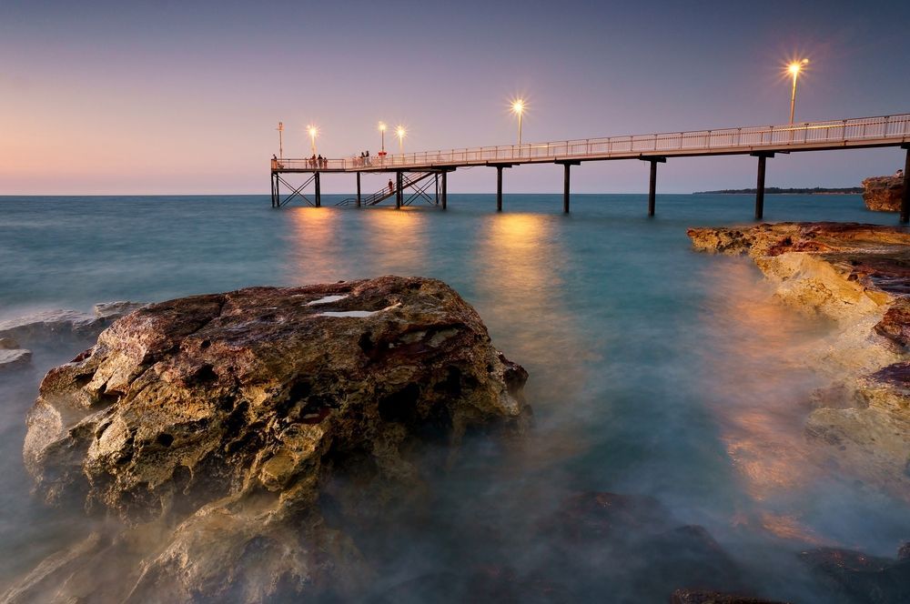 A long pier with glowing lights extends over calm, turquoise water toward a sunset horizon, with rugged rocks in the foreground — Money Spider Conveyancing In Katherine, NT