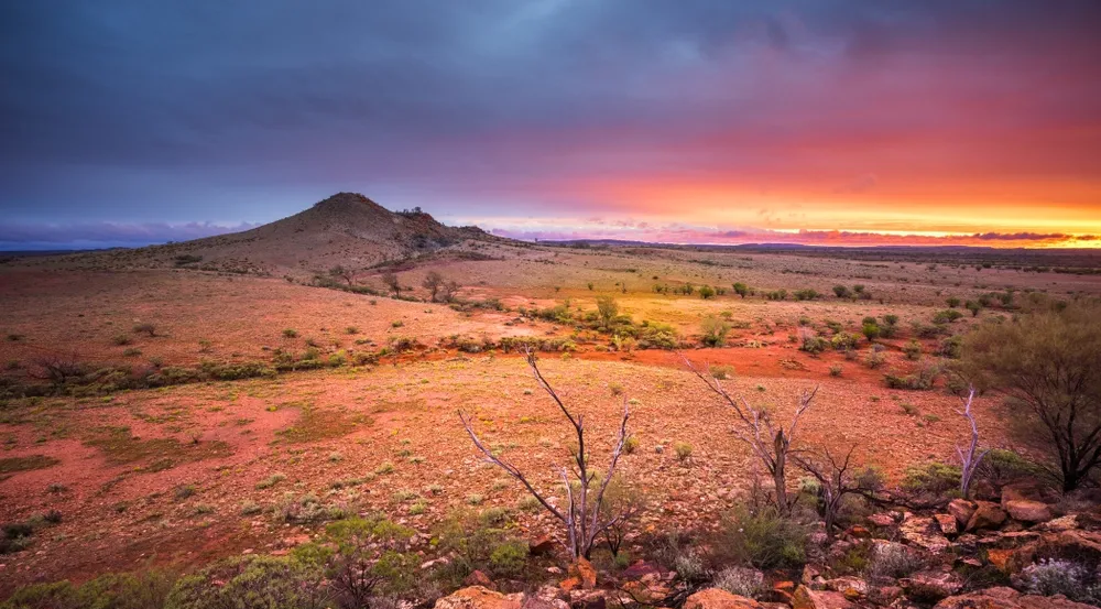 A sunset glows with vibrant orange and purple clouds over a vast, arid desert landscape with a rocky hill in the distance — Money Spider Conveyancing In Katherine, NT