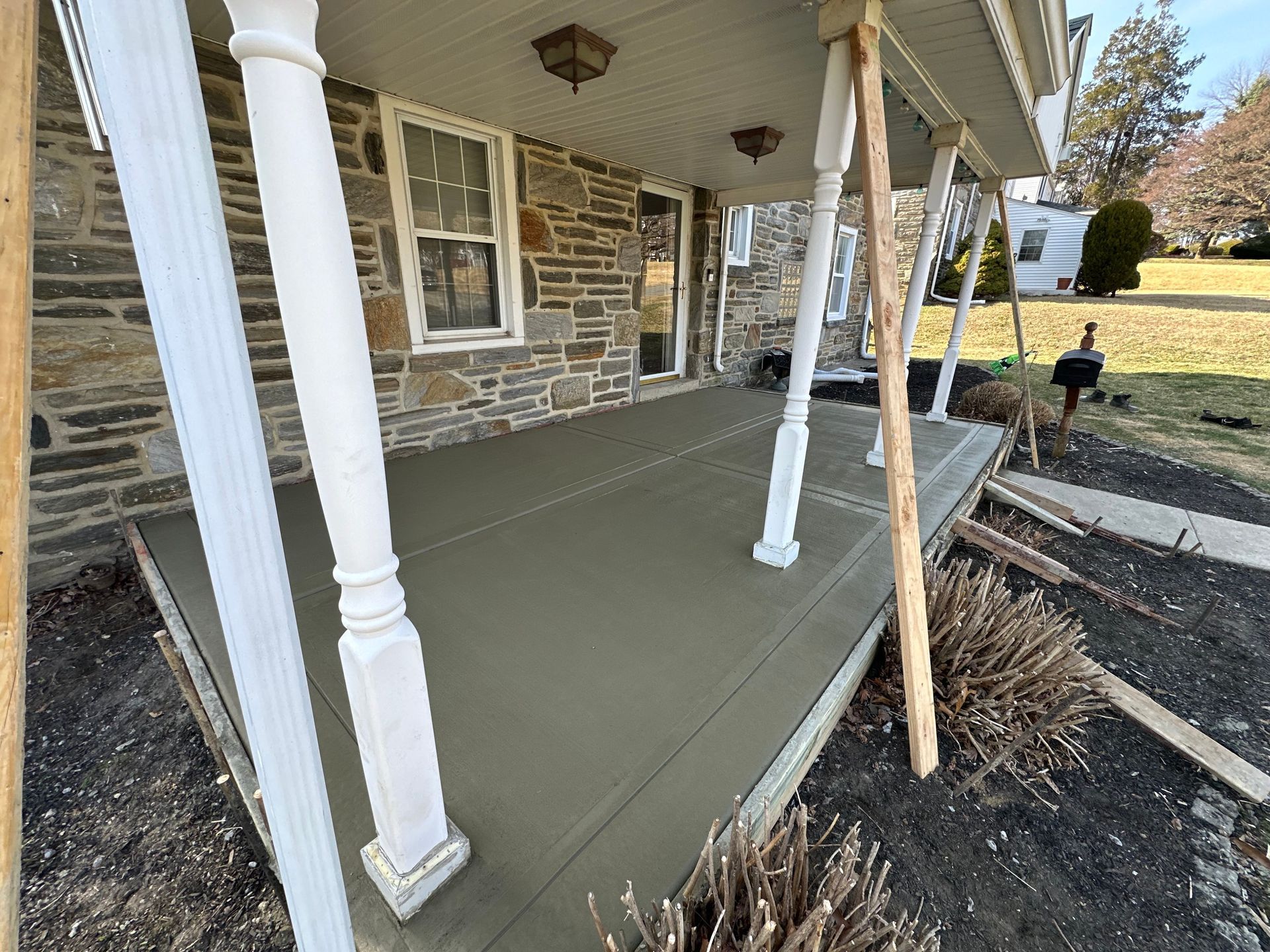 Front porch with fresh gray concrete, white columns, and a stone house exterior under construction.