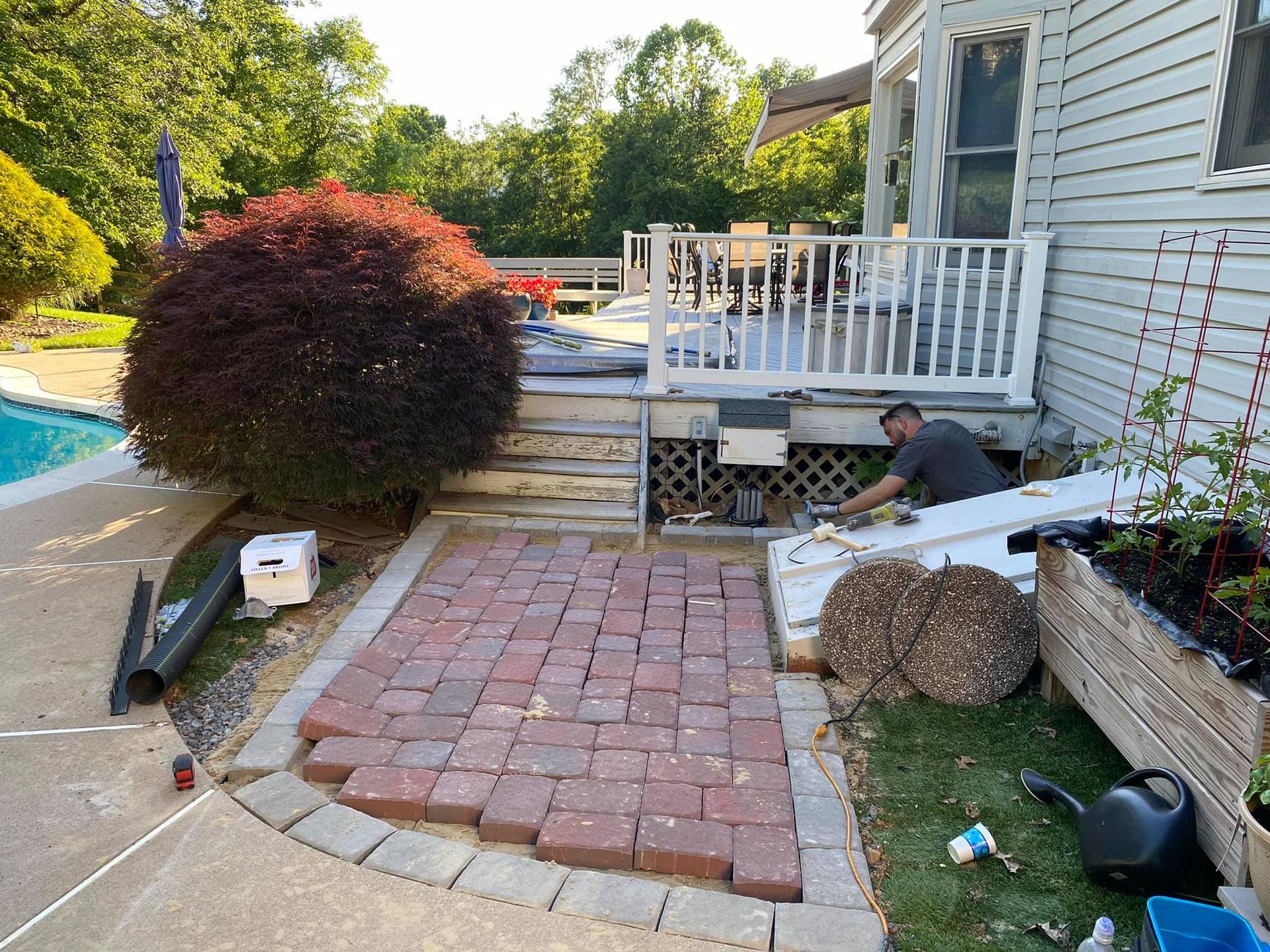 Backyard patio with brick path, pool, and white deck beside a house, with landscaping materials scattered around