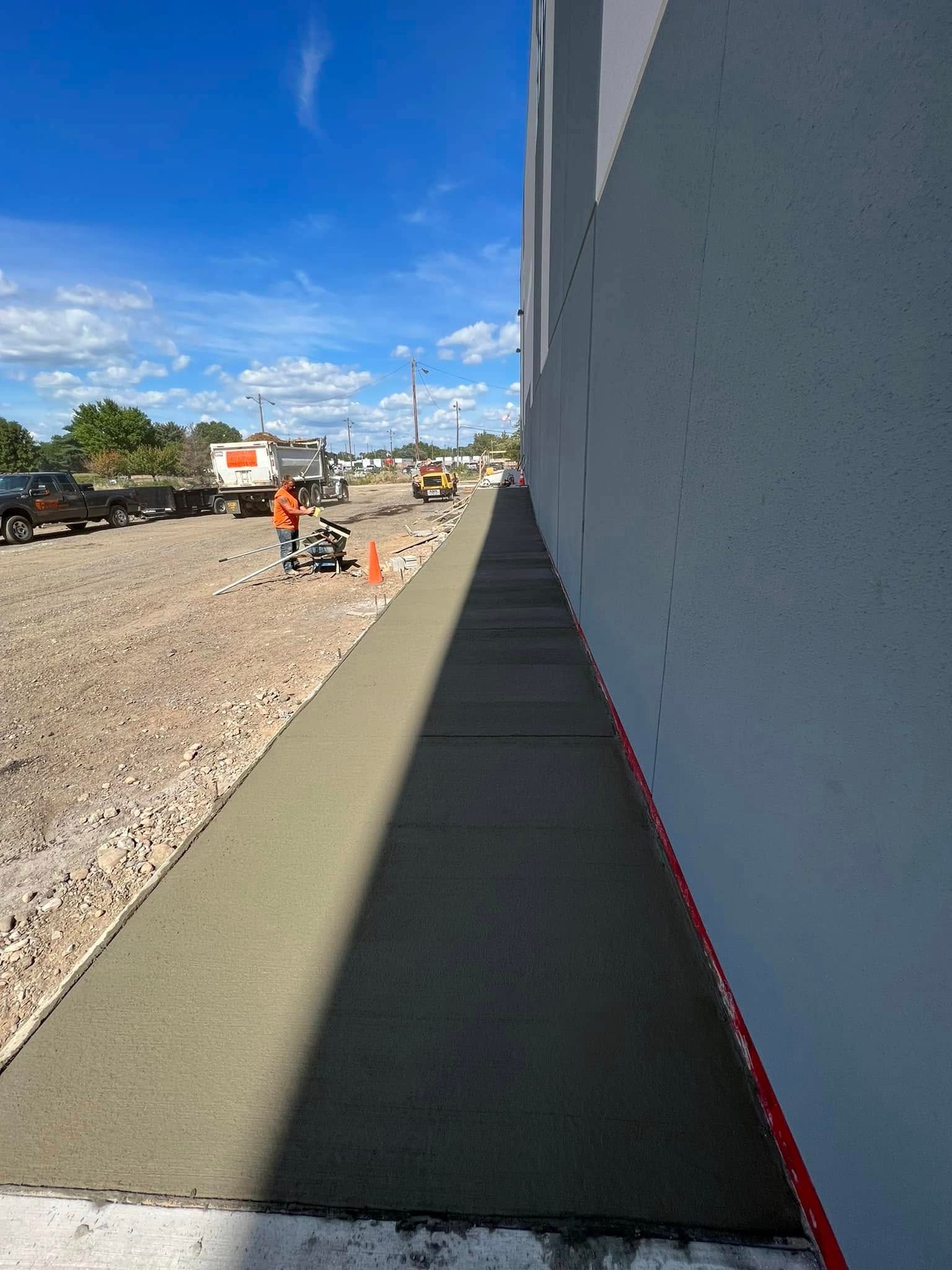 Concrete walkway beside a white building, with orange cones and workers along a gravel construction road under blue sky