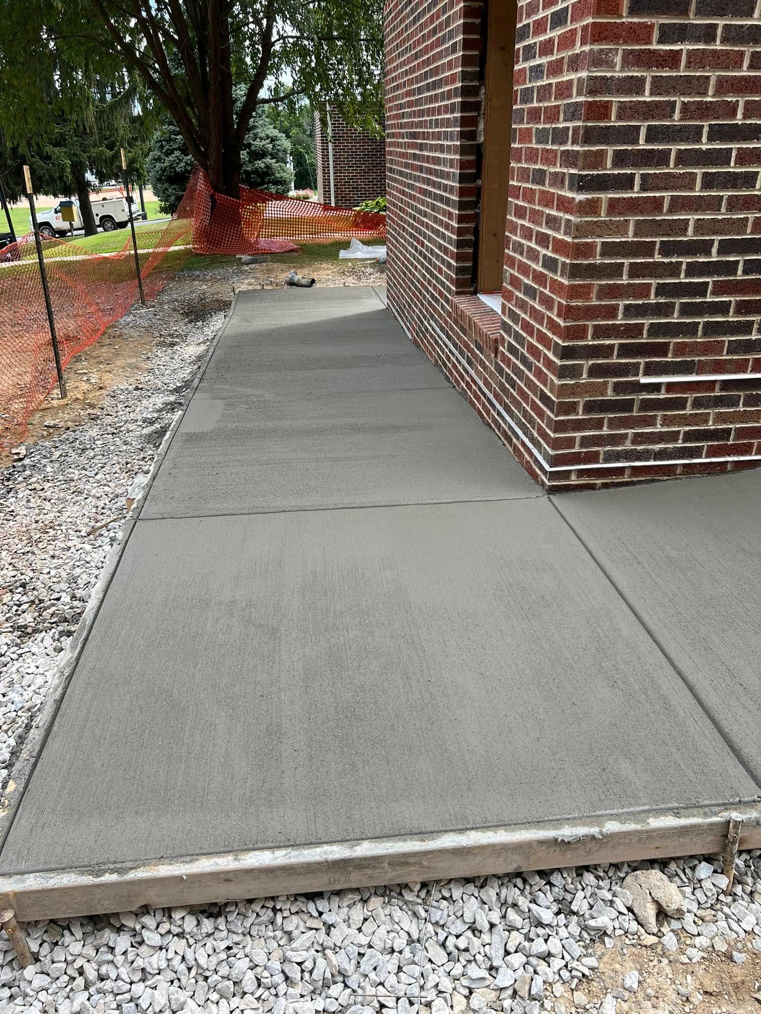 Concrete sidewalk beside a brick building, bordered by gravel and orange construction fencing.