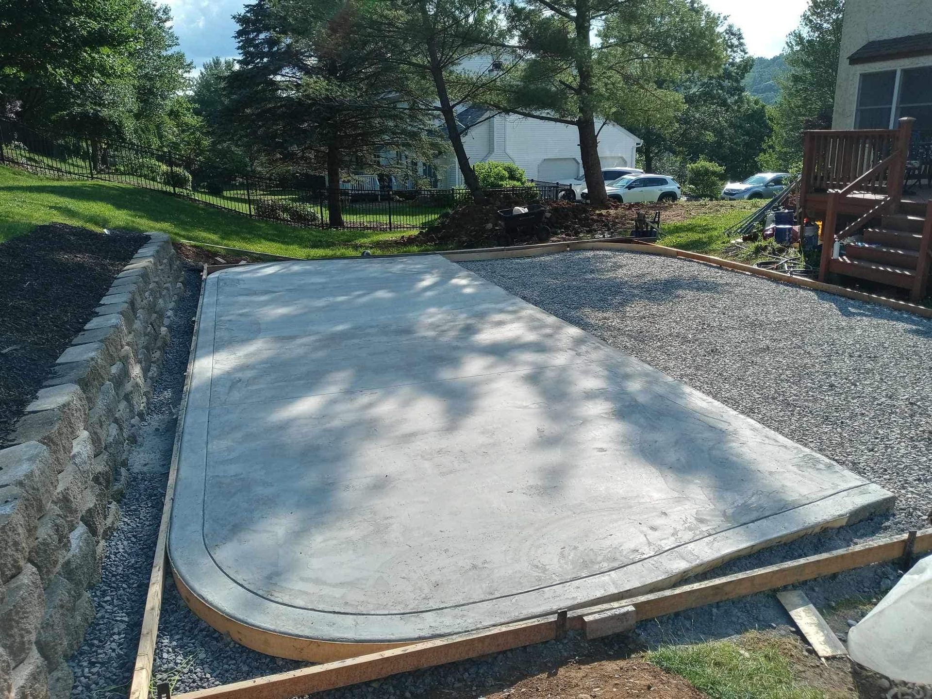 Concrete patio slab in a gravel and stone yard, bordered by trees and a house deck.