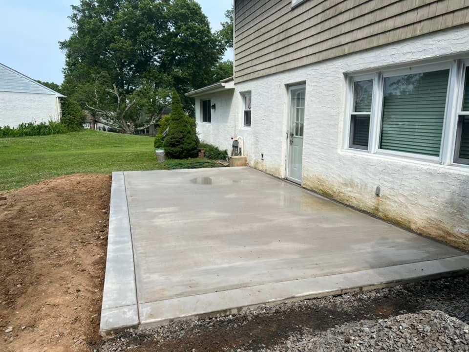 Concrete patio beside a white house with windows, next to a grassy yard and trees