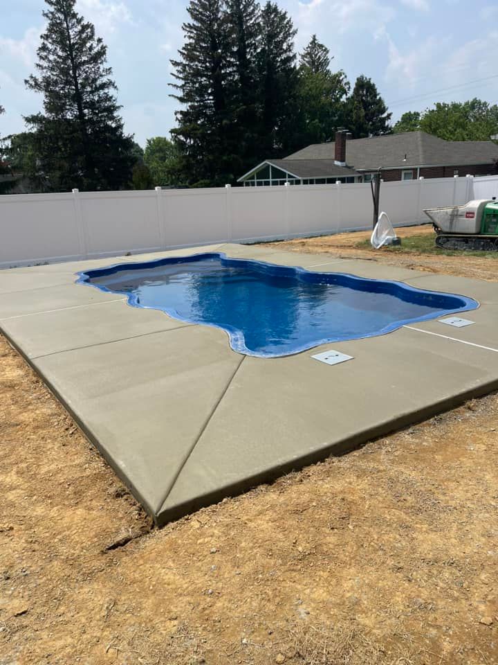 Backyard in-ground pool with blue water and a tan concrete deck, bordered by a white fence and trees