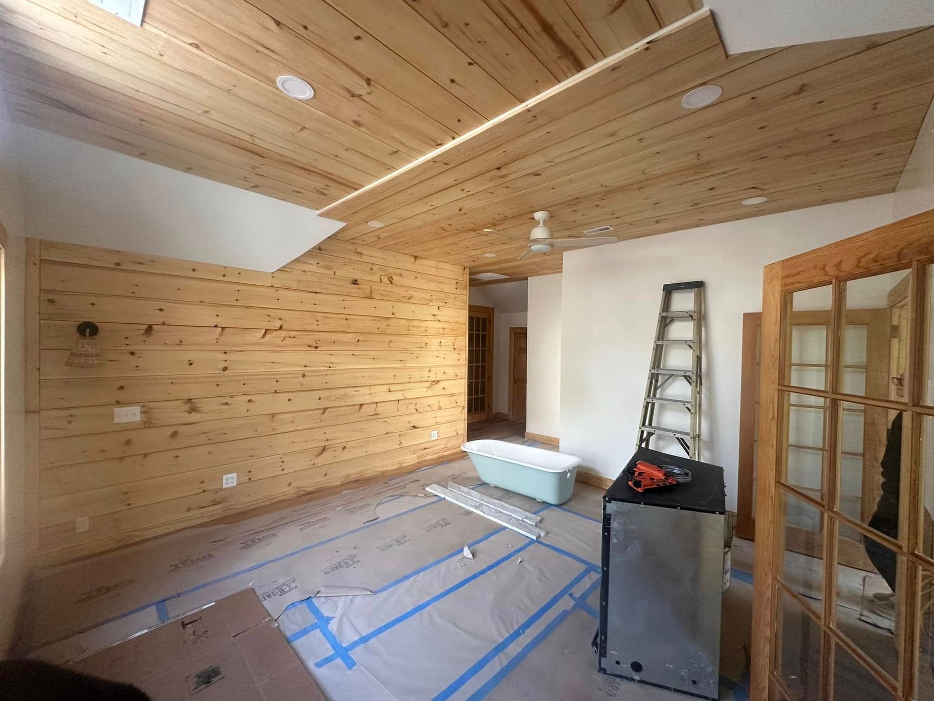 Wood-paneled room with sloped ceiling, ladder, workbench, and taped-off floor under renovation
