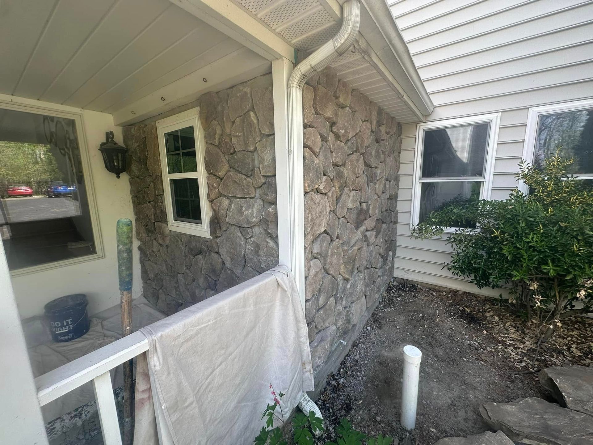 Stone house porch with white railing and window, beside a dirt yard and shrub
