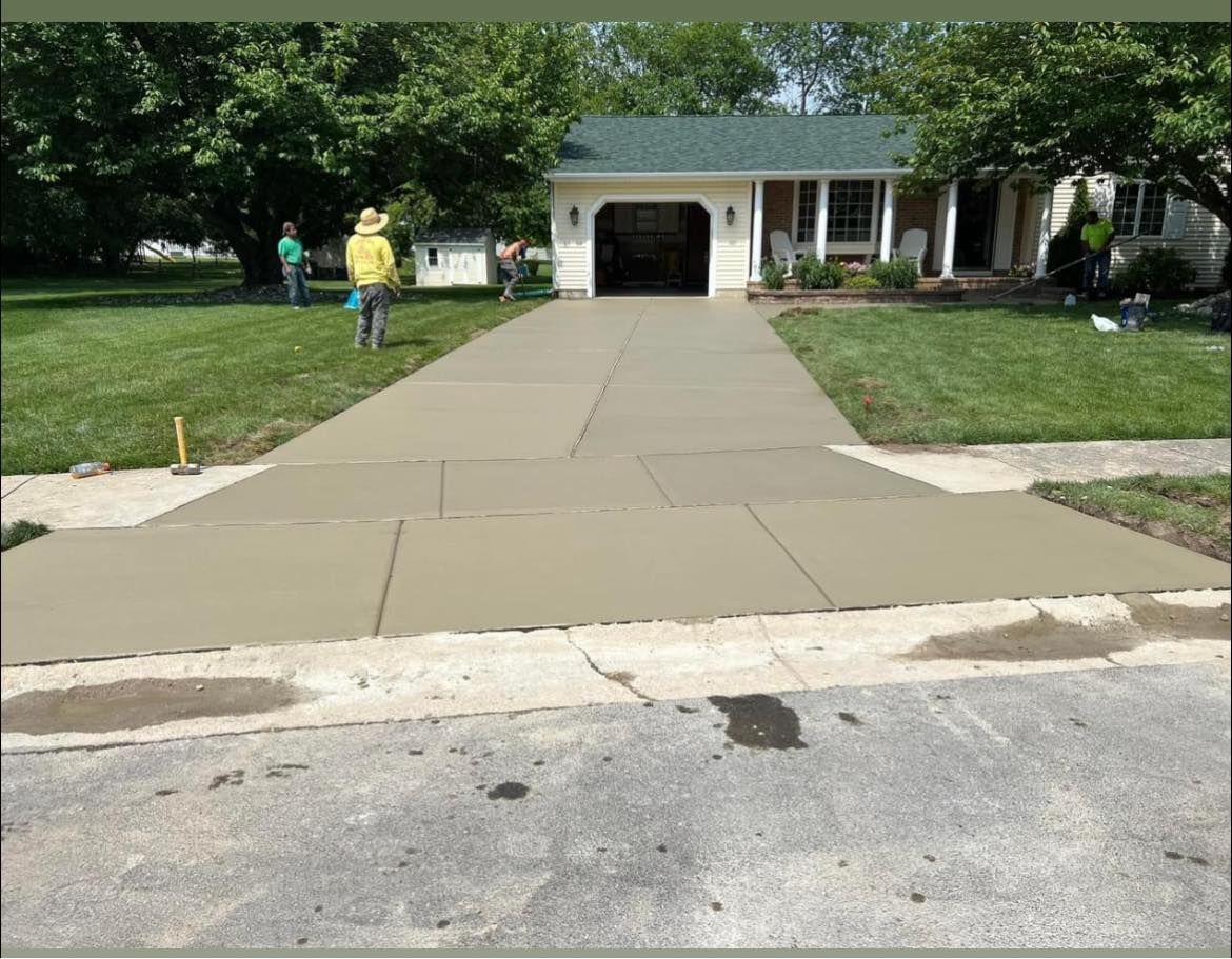 Wide sidewalk leading to a house garage, with two people on the lawn and green trees nearby.