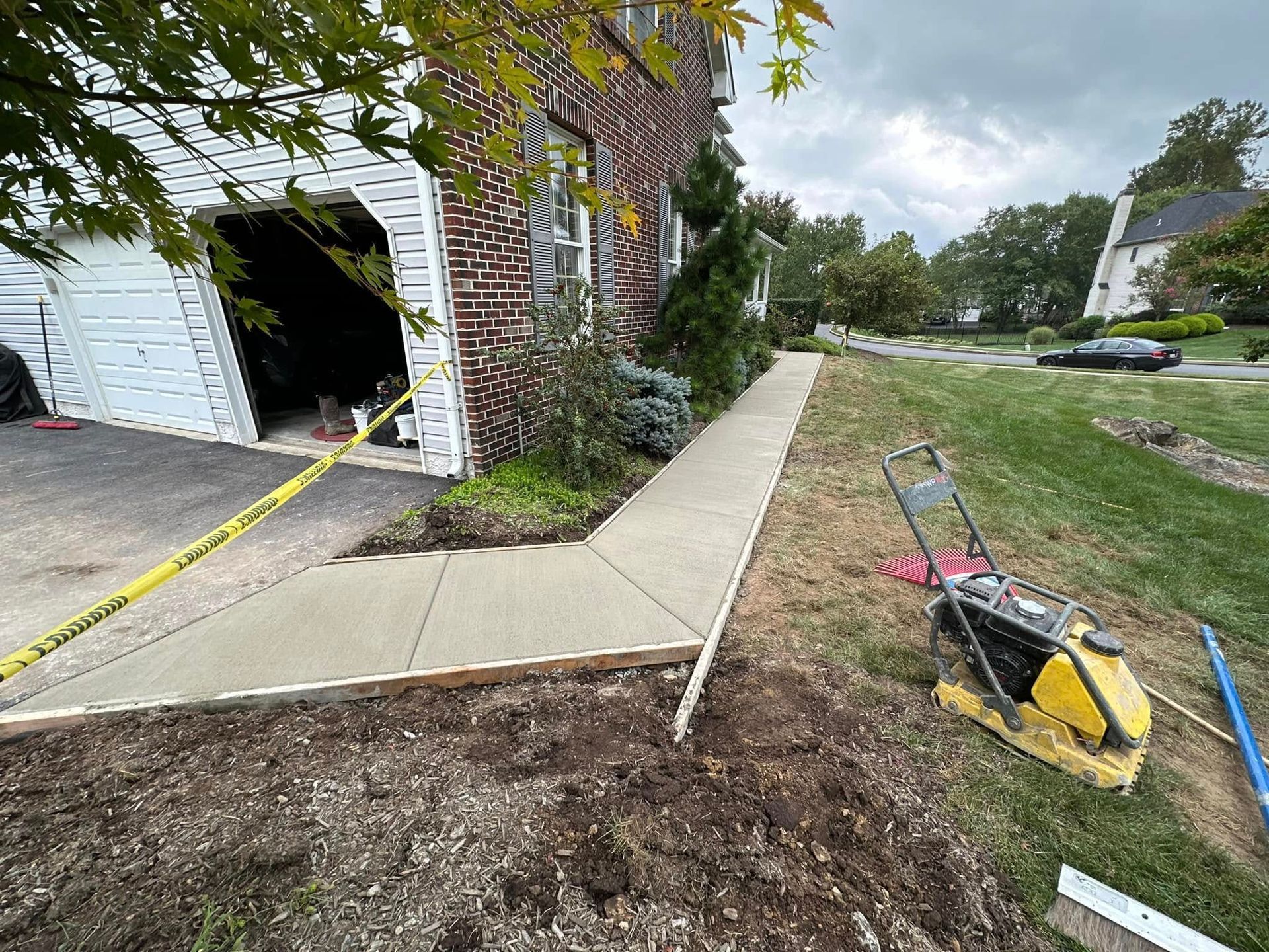 Damaged concrete walkway beside a brick house, with a tipped wheelbarrow and dirt piles near a garage.