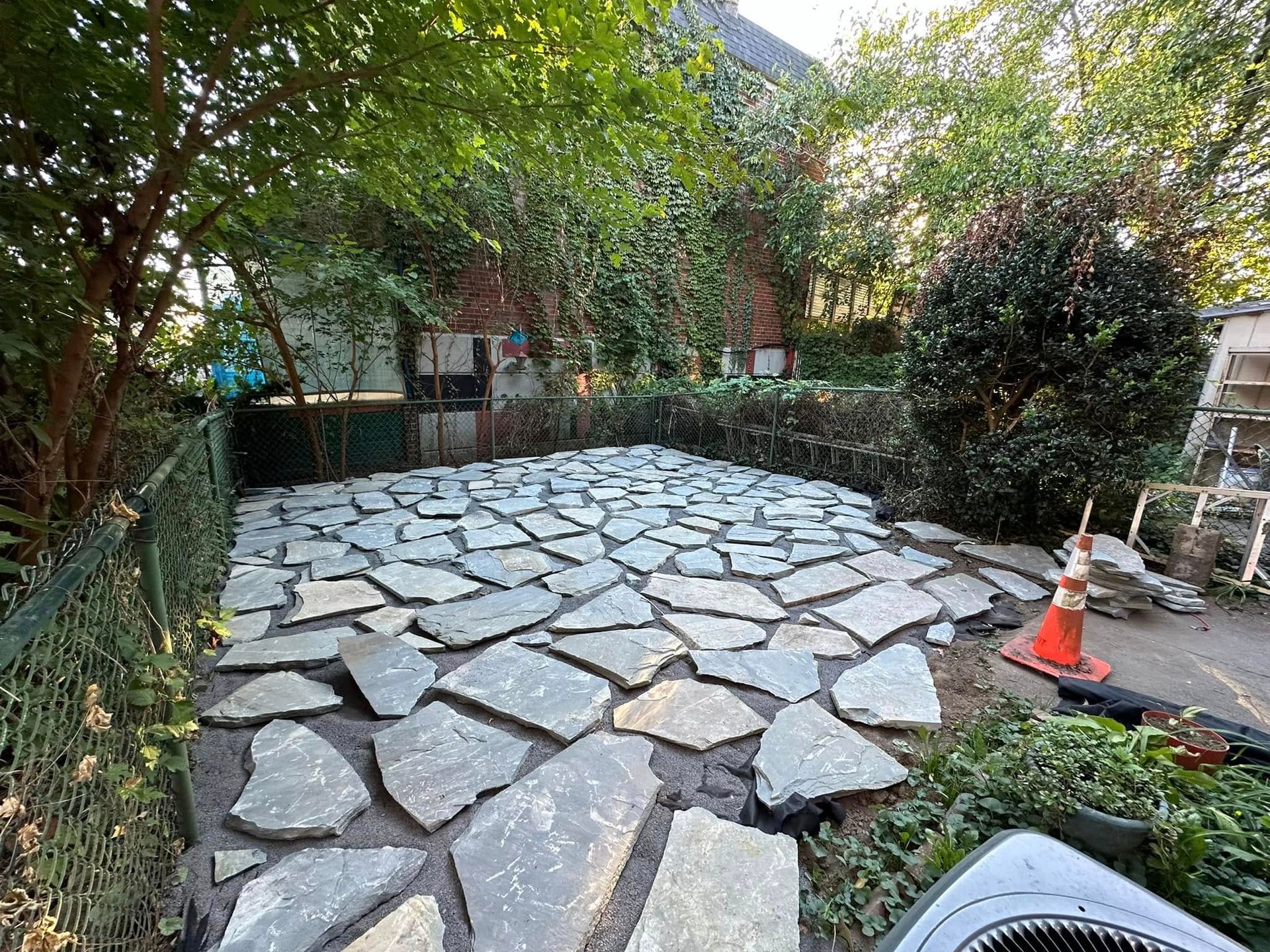 Stone-paved garden courtyard with trees, fence, and an orange traffic cone on the right