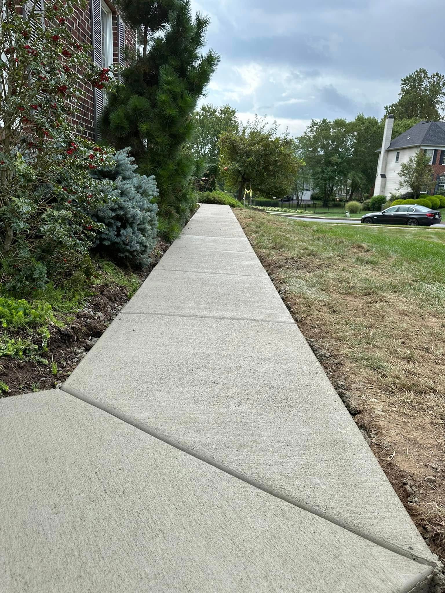 Concrete sidewalk beside bushes and grass in a suburban neighborhood under cloudy skies.