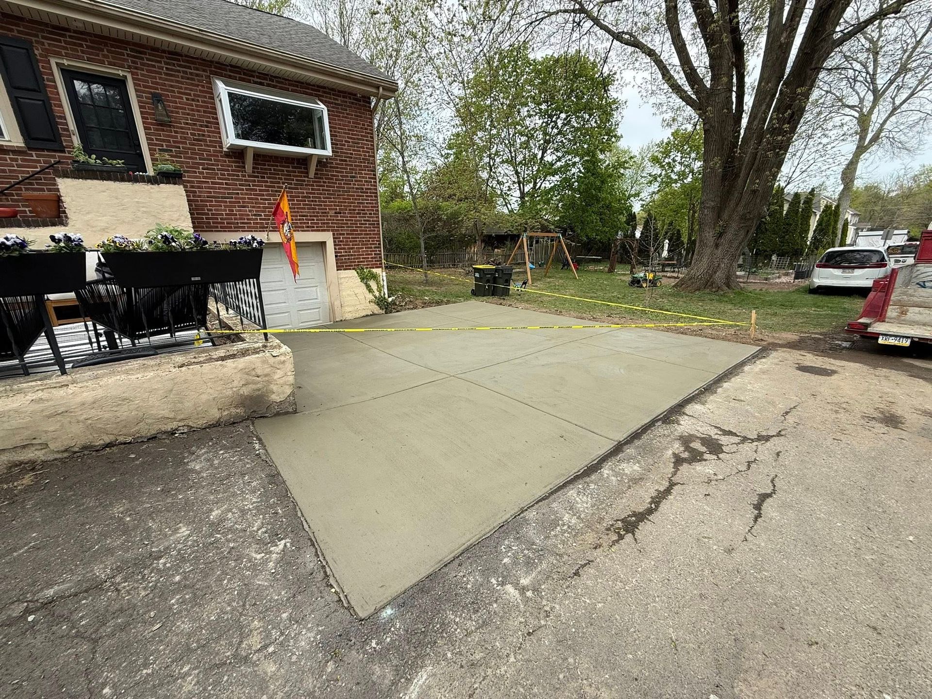 Concrete driveway and patio beside a brick house, with a tree-lined yard and parked car nearby.