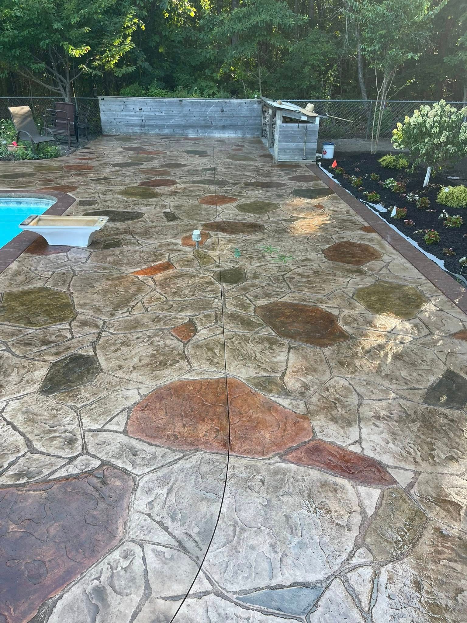 Backyard stone patio with patches and a small pool at left, bordered by greenery and a low wall.