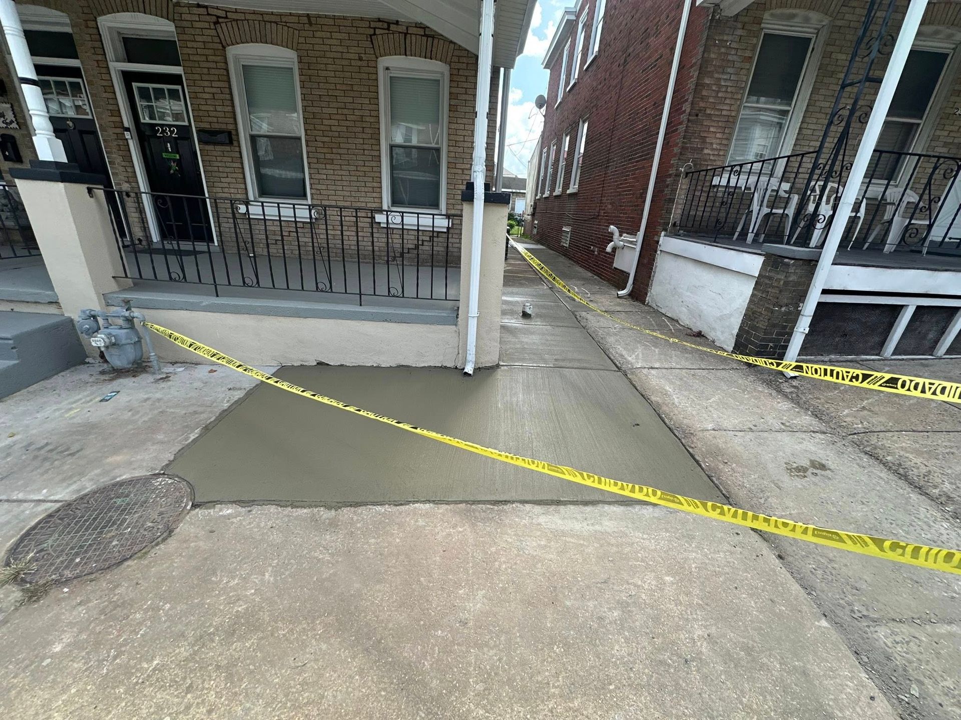 Caution tape blocks a concrete walkway between brick buildings with a damaged, patched section.