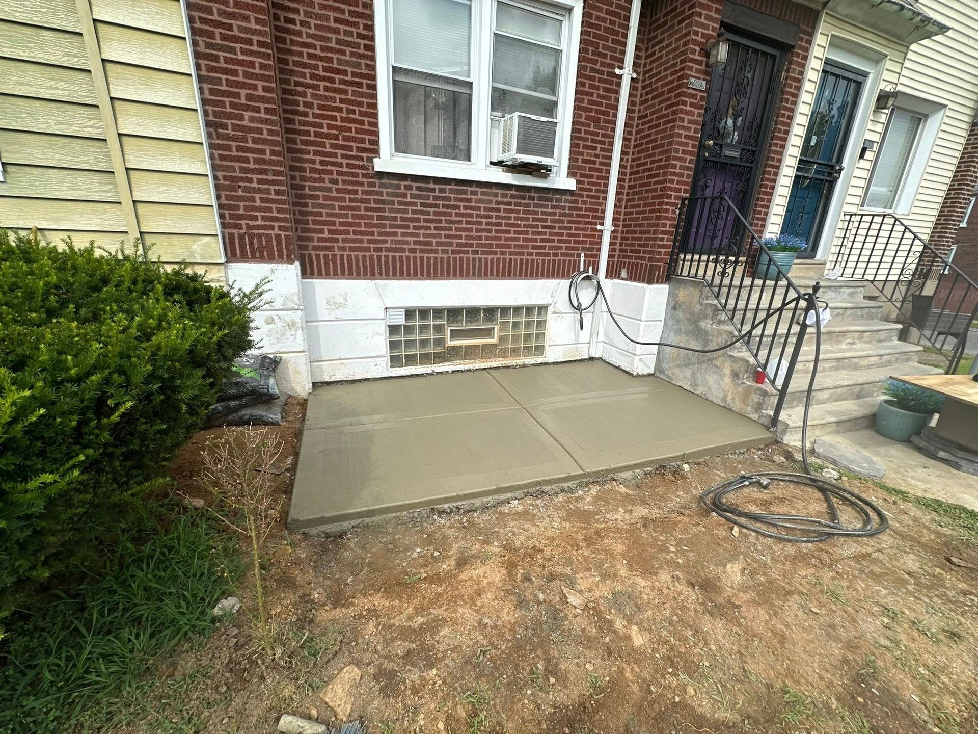 Fresh concrete patio slab beside a brick house and front steps, with a shrub and unfinished dirt yard