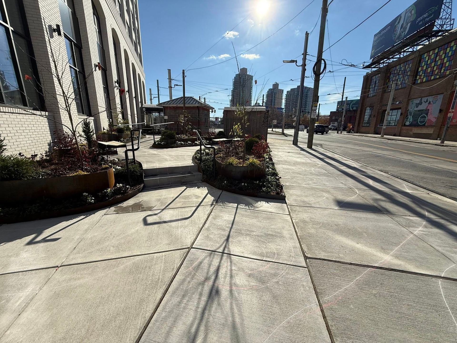 Sunlit city sidewalk beside tram tracks, planter boxes, and buildings under a clear blue sky.