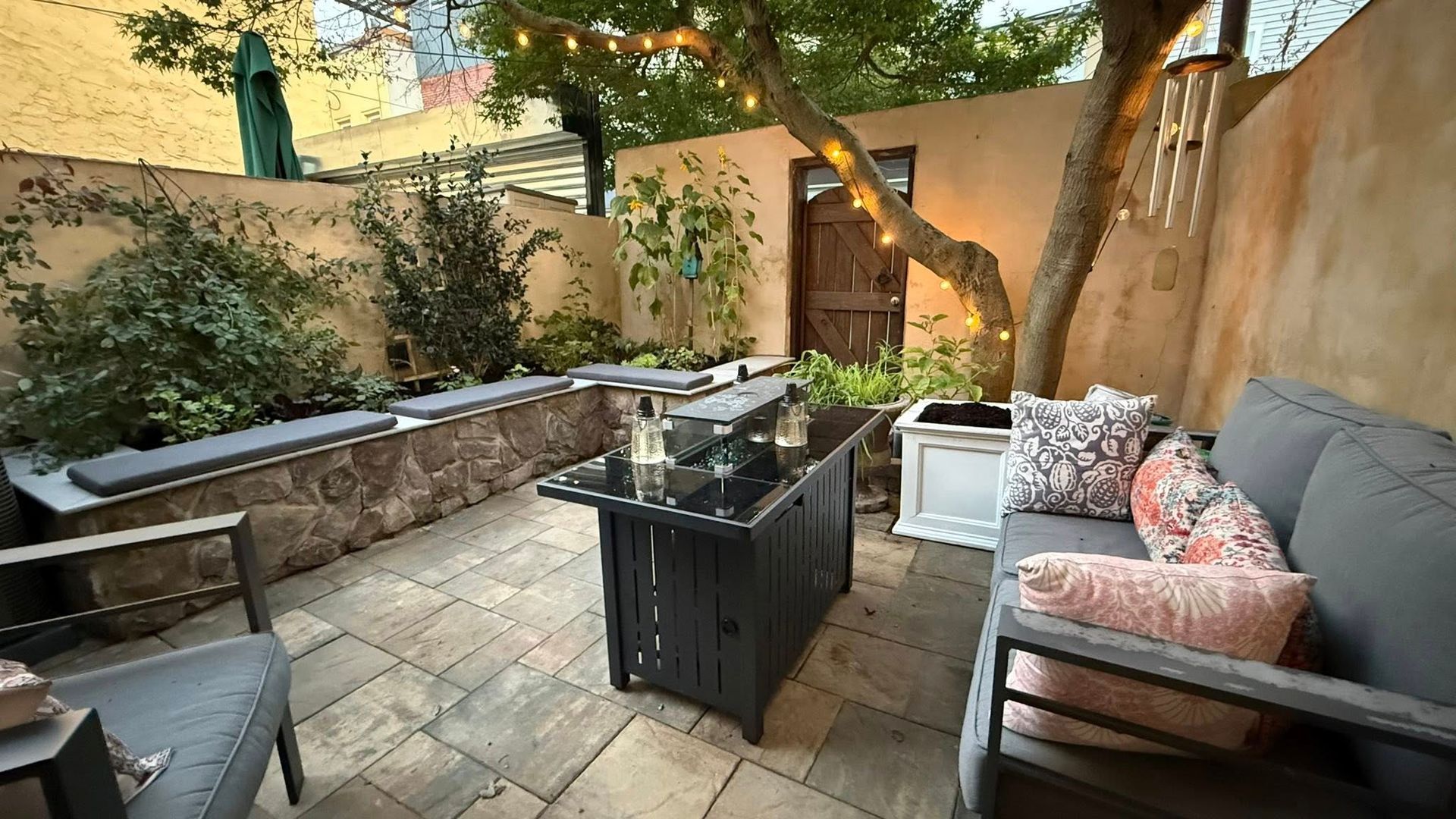 Cozy backyard patio with seating, fire pit table, string lights, and potted plants under trees.
