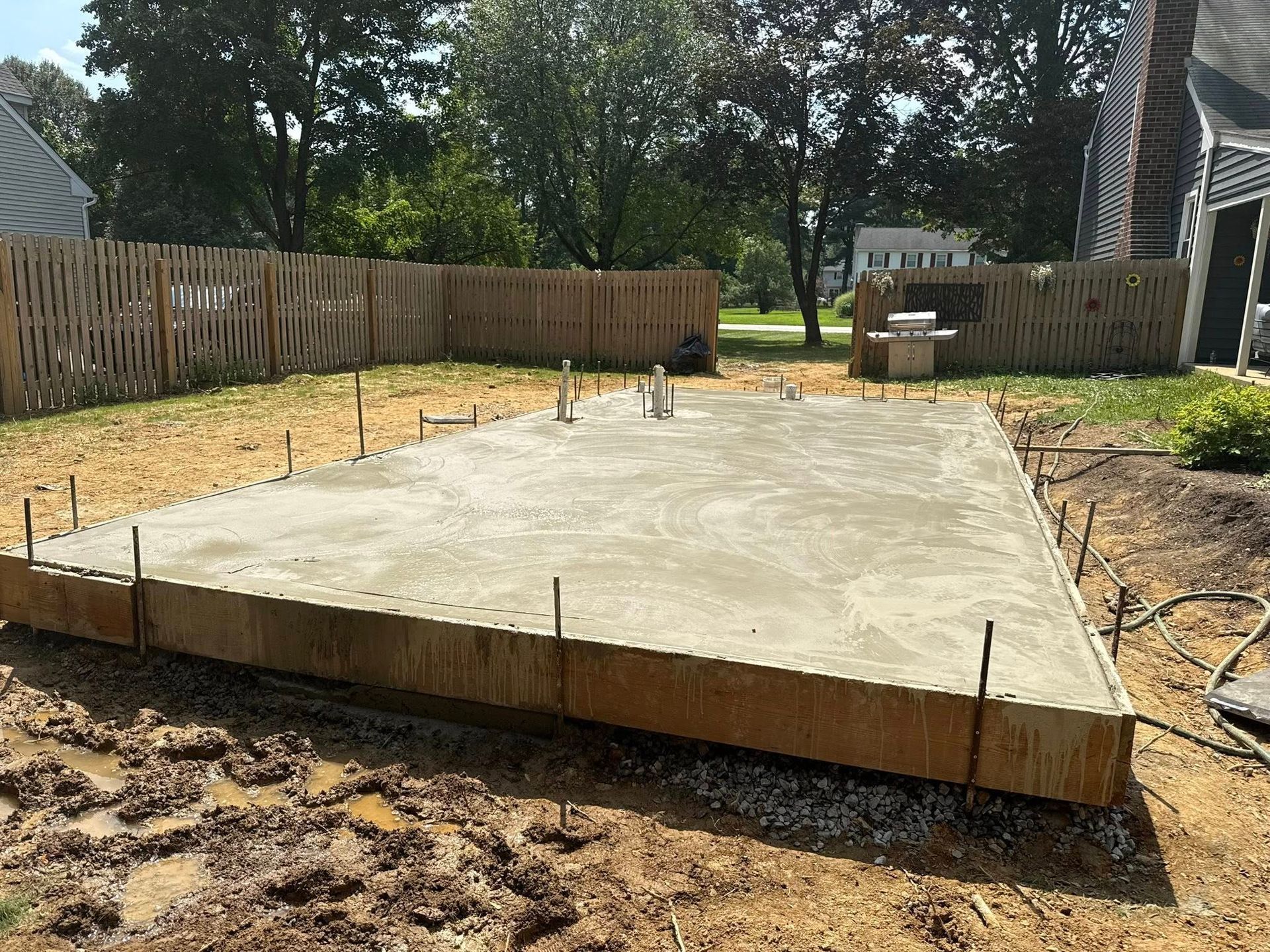 Concrete slab foundation in a fenced backyard with dirt, trees, and a house in the background