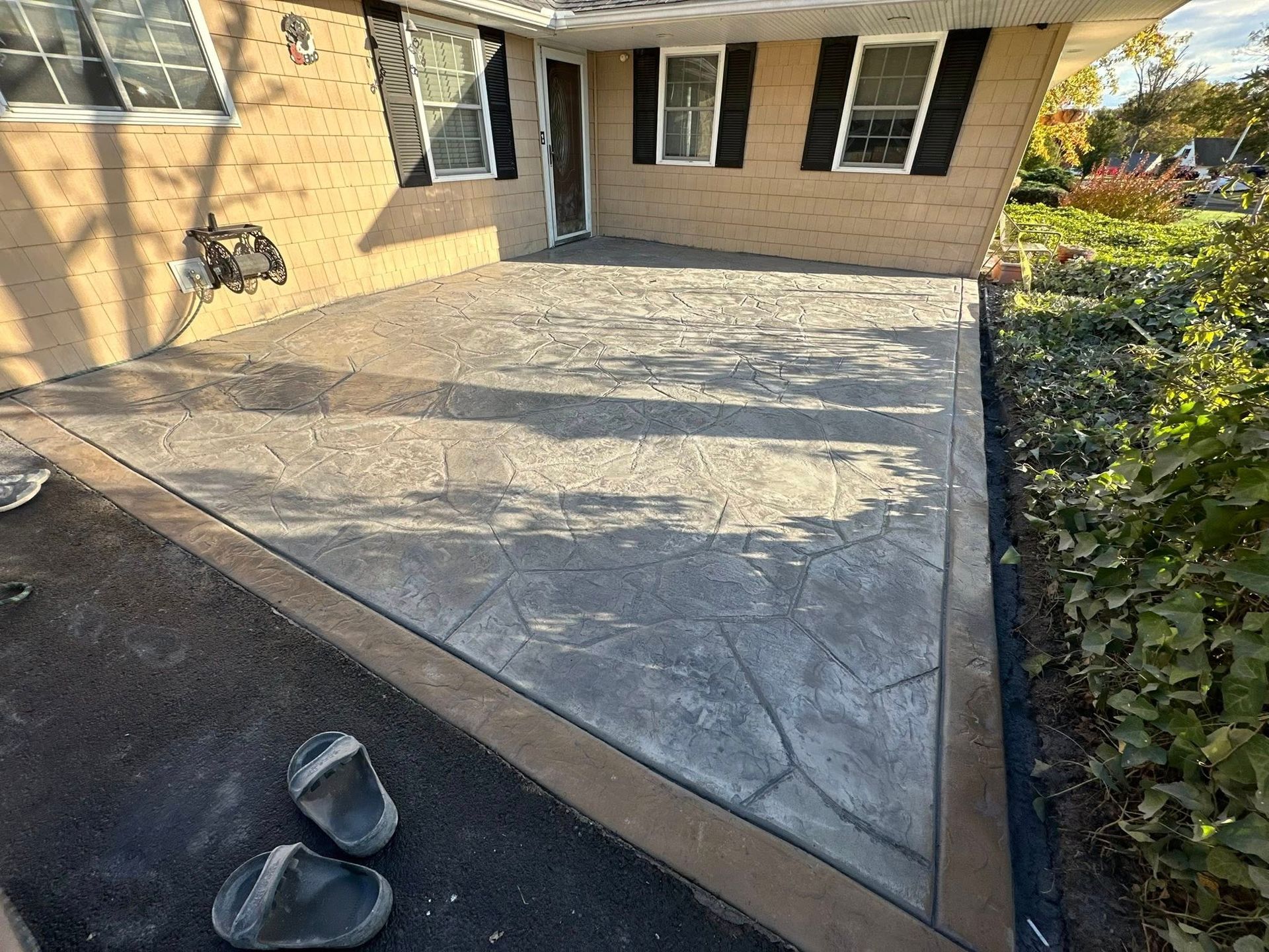 Concrete patio beside a beige house, with a pair of shoes at the edge and hedge along the right side.