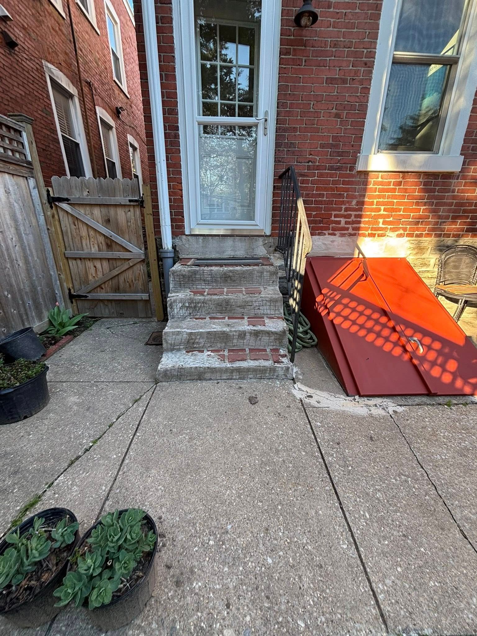 Brick house entrance with worn steps, side door, and red basement hatch in a narrow alley courtyard