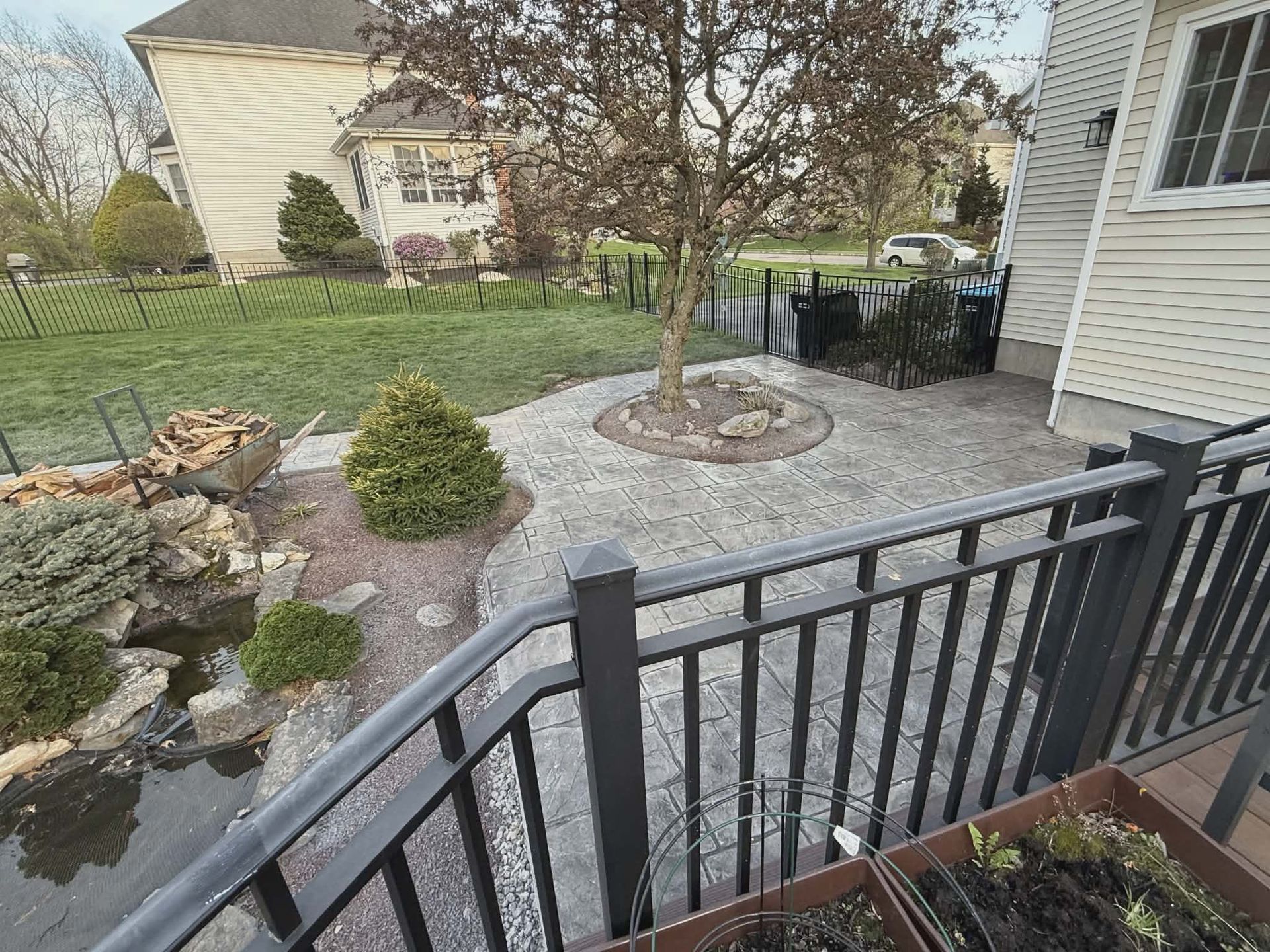 Front yard patio with stone pavers, small tree, shrubs, and a black metal railing by a house