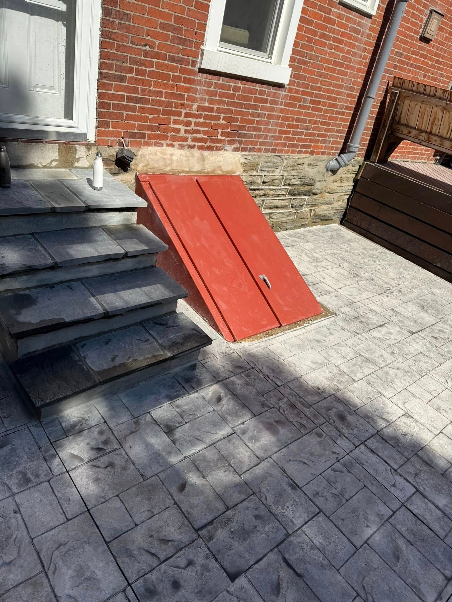 Red cellar hatch beside brick house wall, next to stone steps and paved patio.
