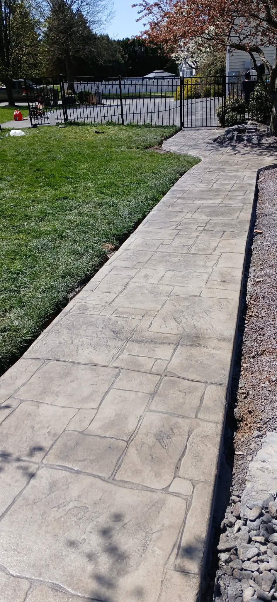 Curved concrete path beside a grassy lawn and gravel edge, leading toward a white fence and house.
