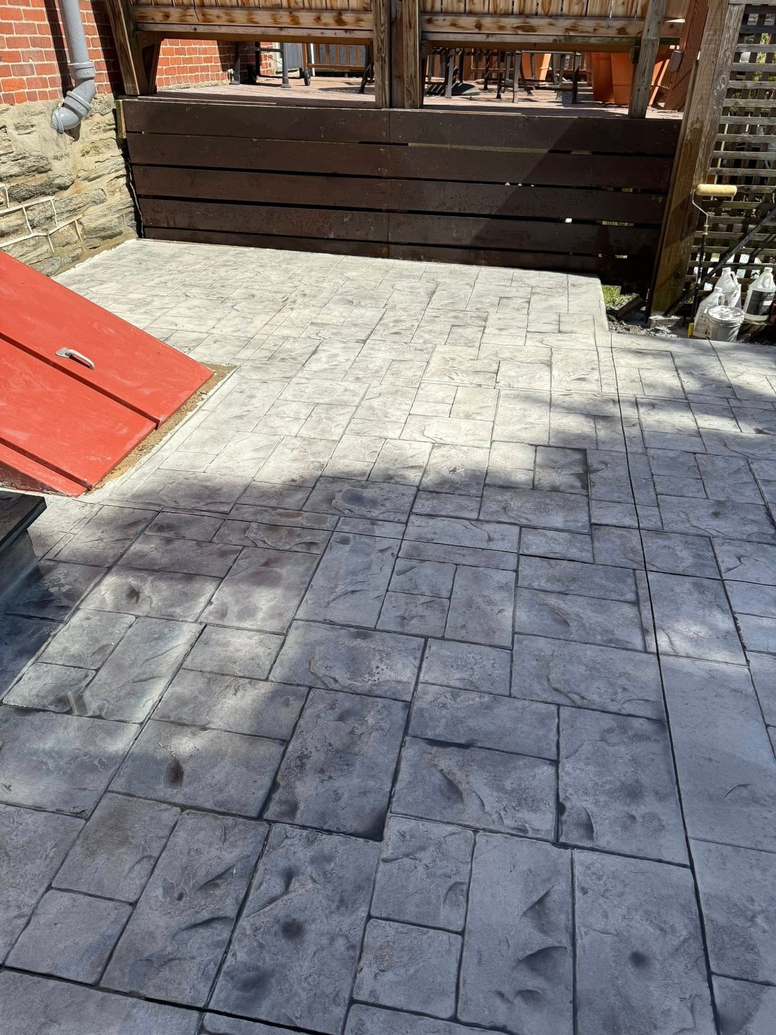 Stone patio in a yard beside a wooden deck and a red ramp, with sunlight and shadows on the ground