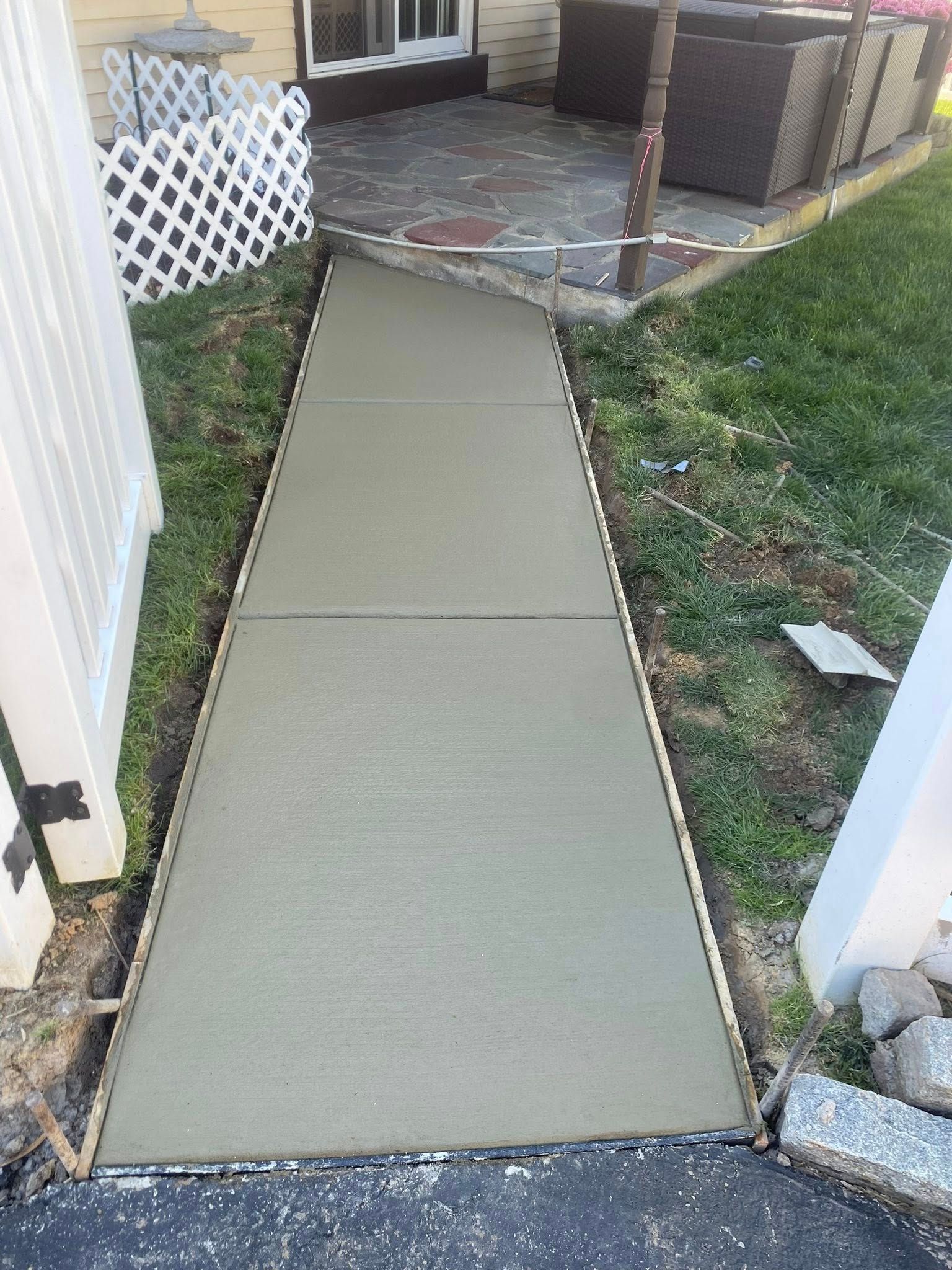 Fresh concrete walkway leading to a fenced backyard patio between a white fence and a house.