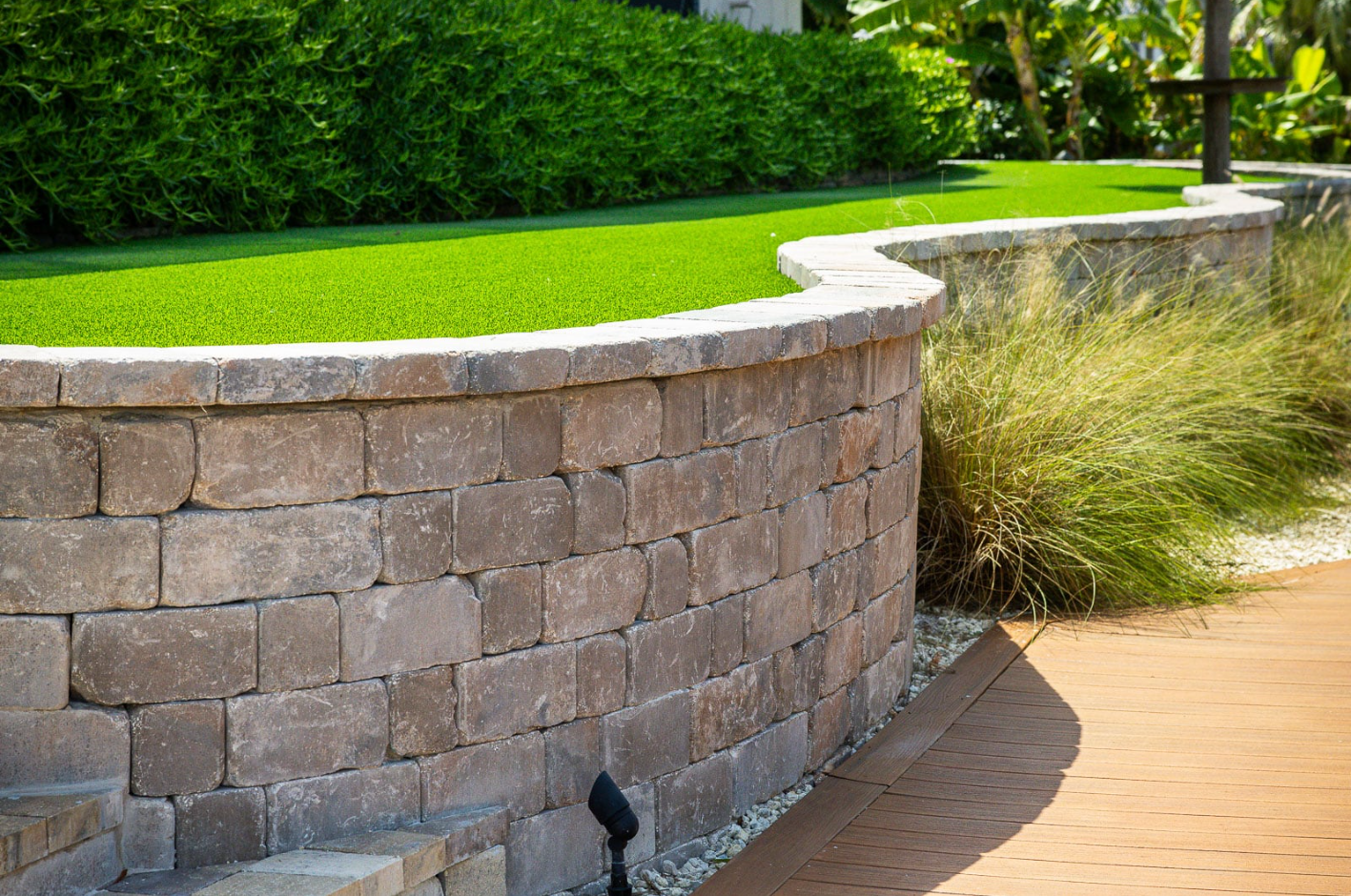 Curved stone retaining wall edging a green lawn beside a tan paved walkway