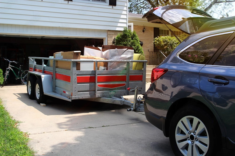 A car hitched to a trailer loaded with moving boxes, parked on a driveway in front of a house.