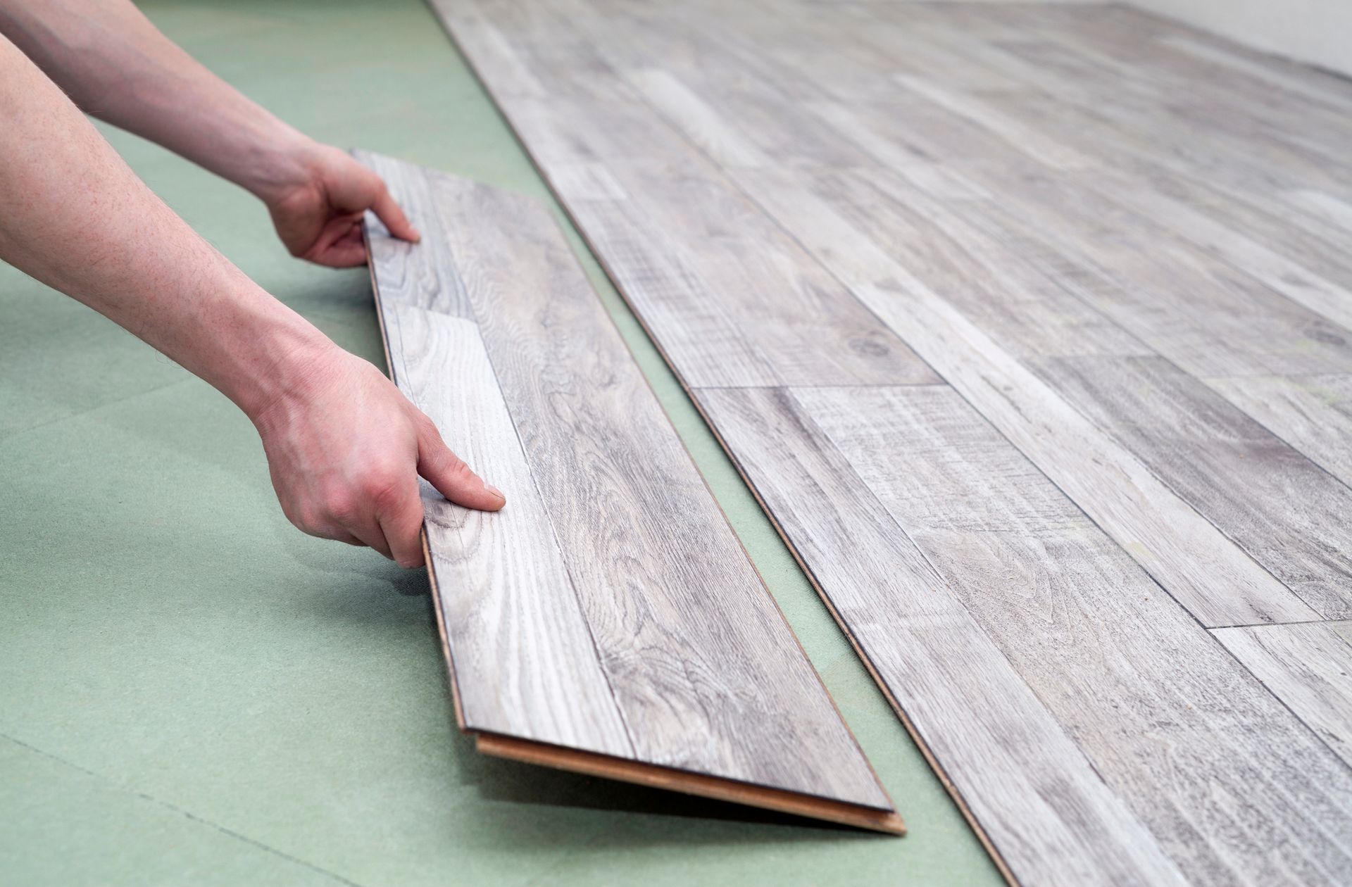 Mani che installano un pavimento in legno su un sottofondo verde.