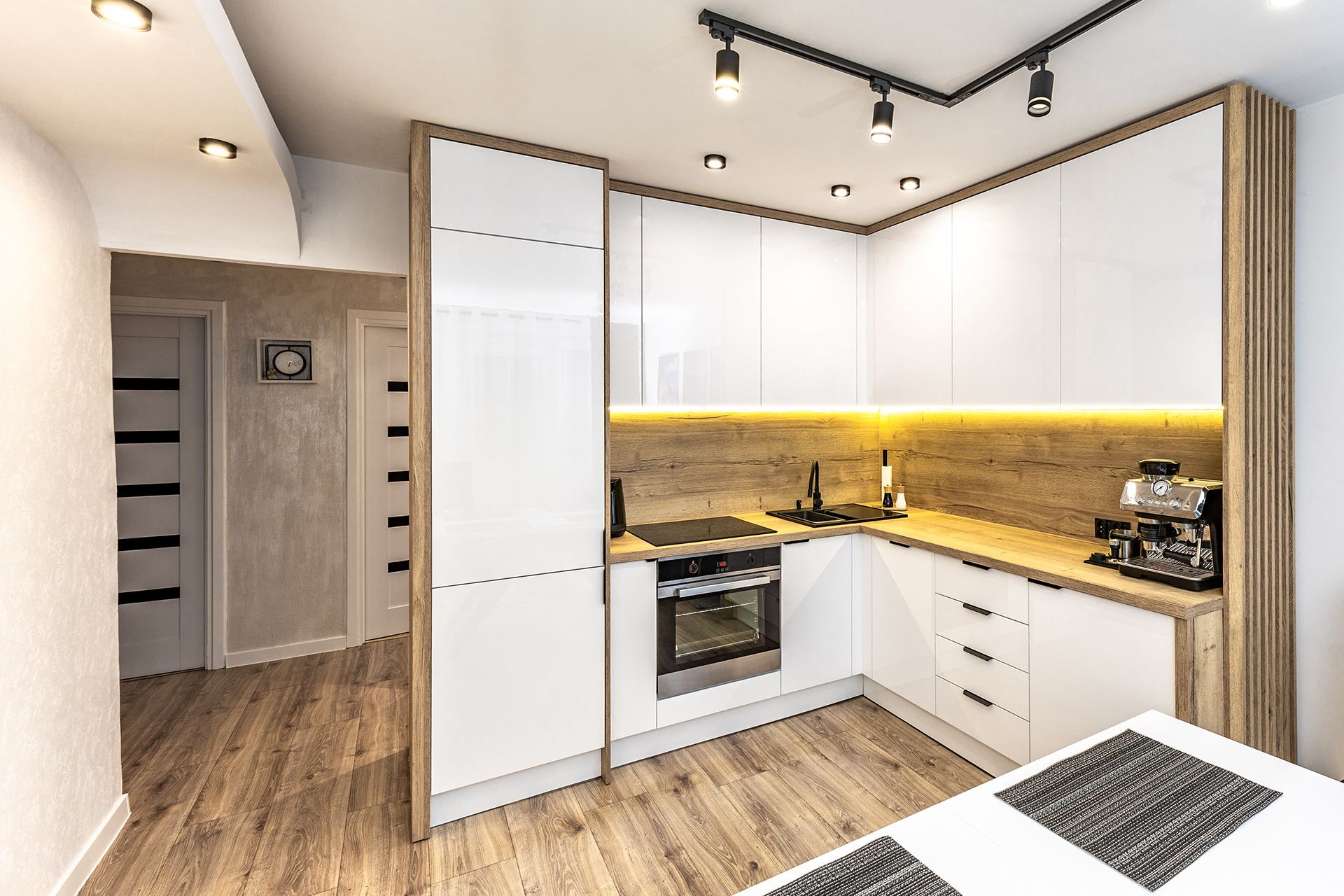 Modern L-shaped kitchen with white cabinets, wood-tone counters and backsplash, under-cabinet lighting, and track lights.