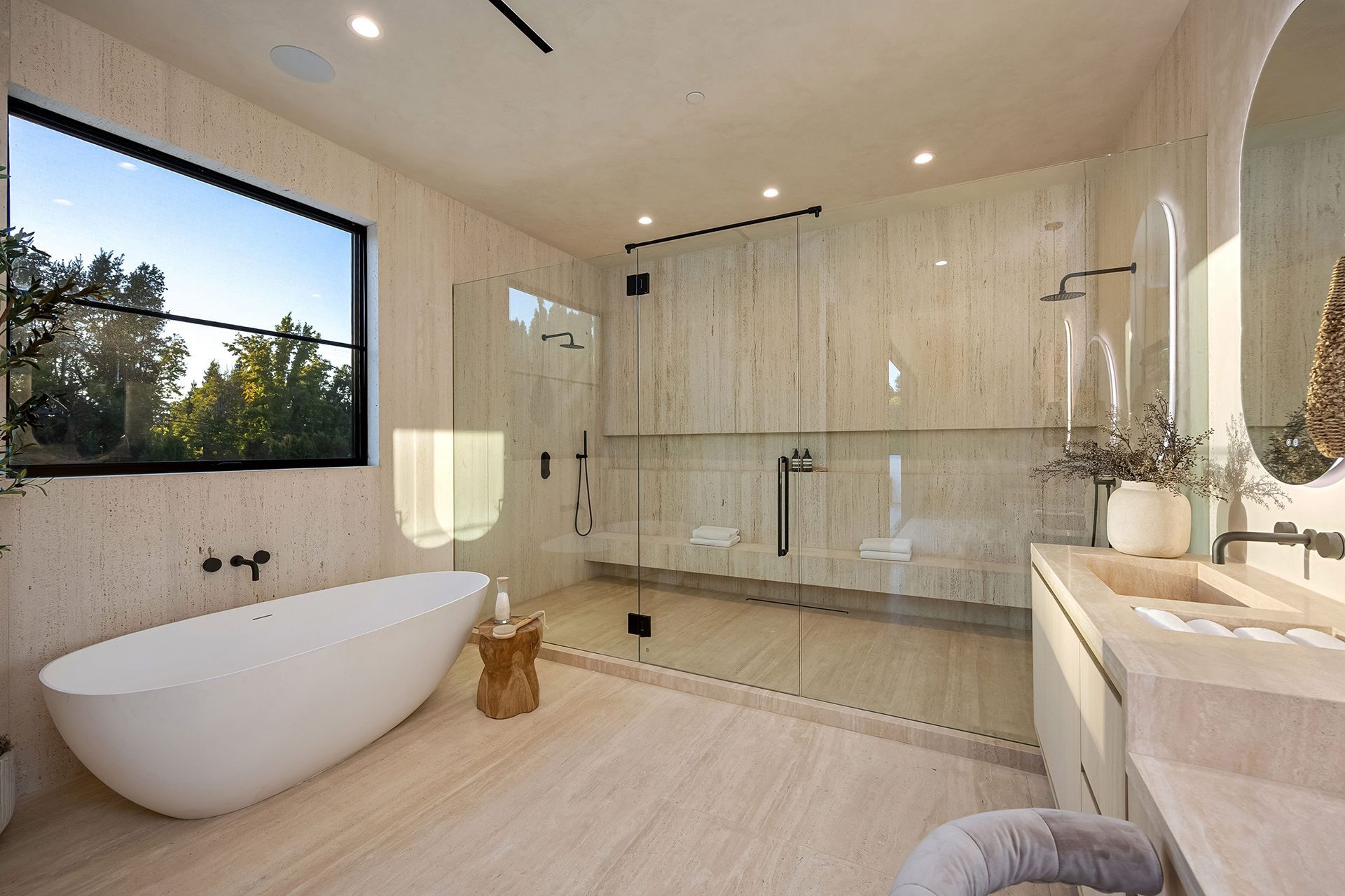 A modern, light-toned bathroom featuring a standalone white oval tub, a spacious glass-enclosed shower, and stone walls.