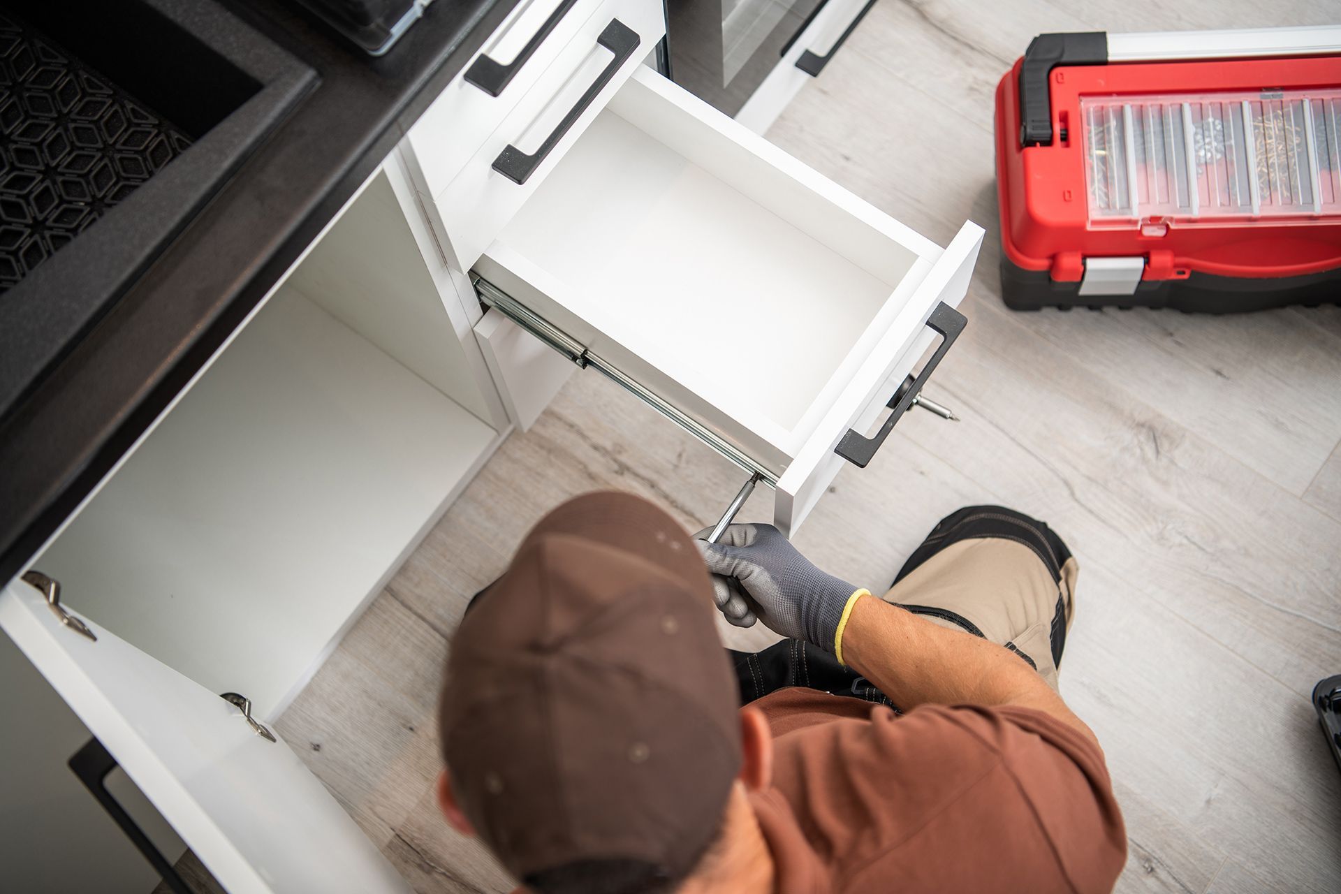 A worker wearing a brown cap and gloves installs a drawer in a kitchen cabinet next to a red toolbox on the floor.