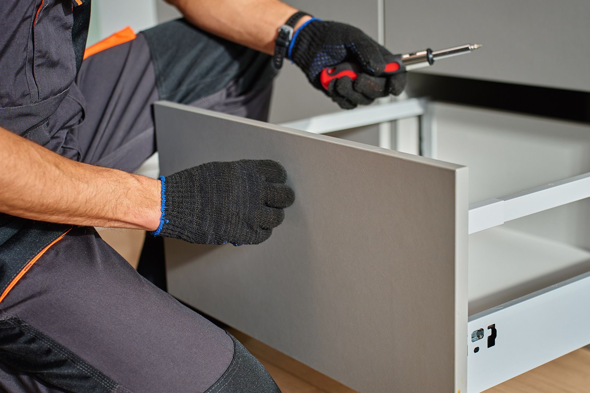 A person in work clothes and gloves uses a screwdriver to assemble a grey furniture drawer.
