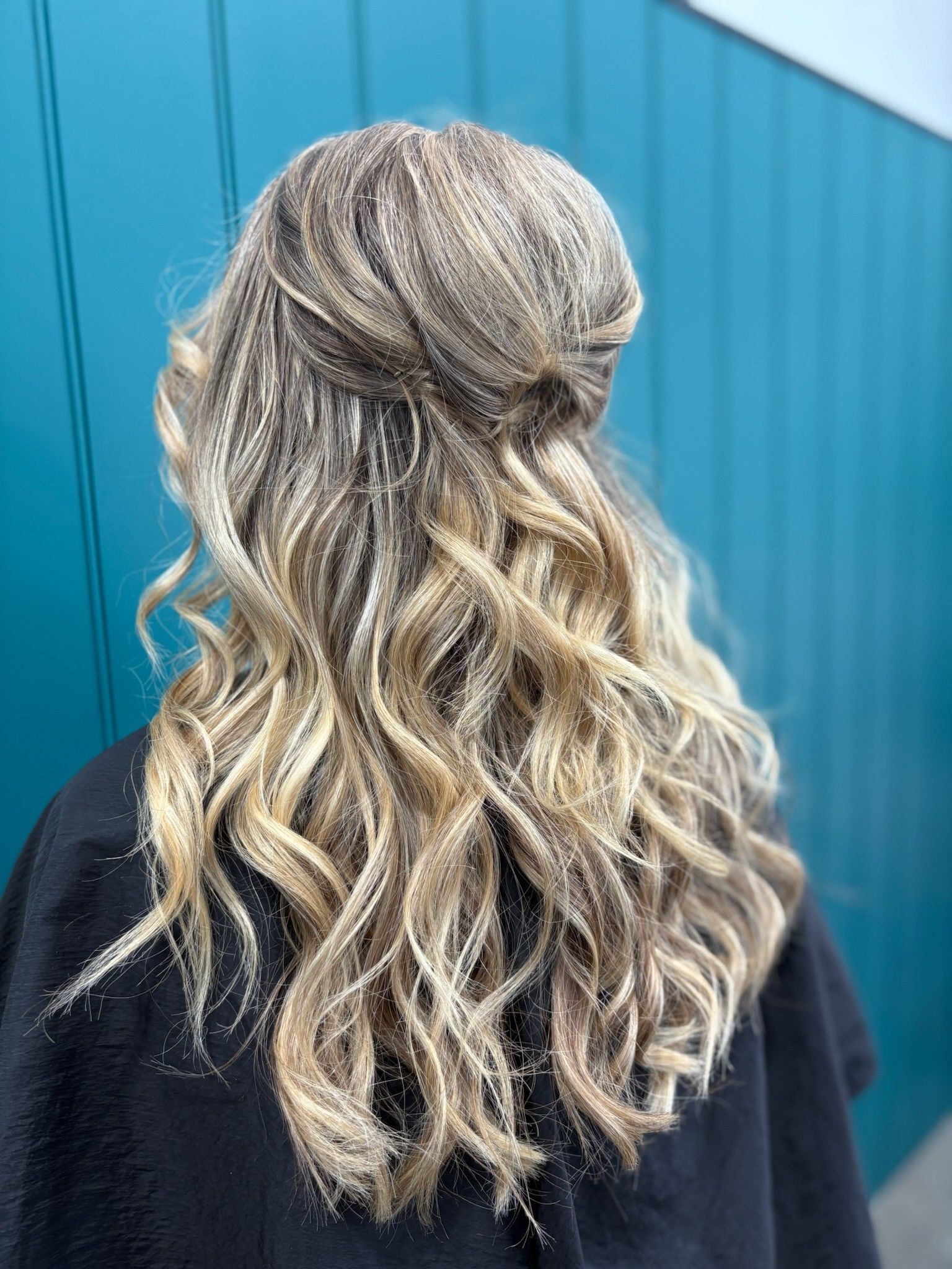 Wavy blonde hair with darker roots, viewed from behind against a teal wall