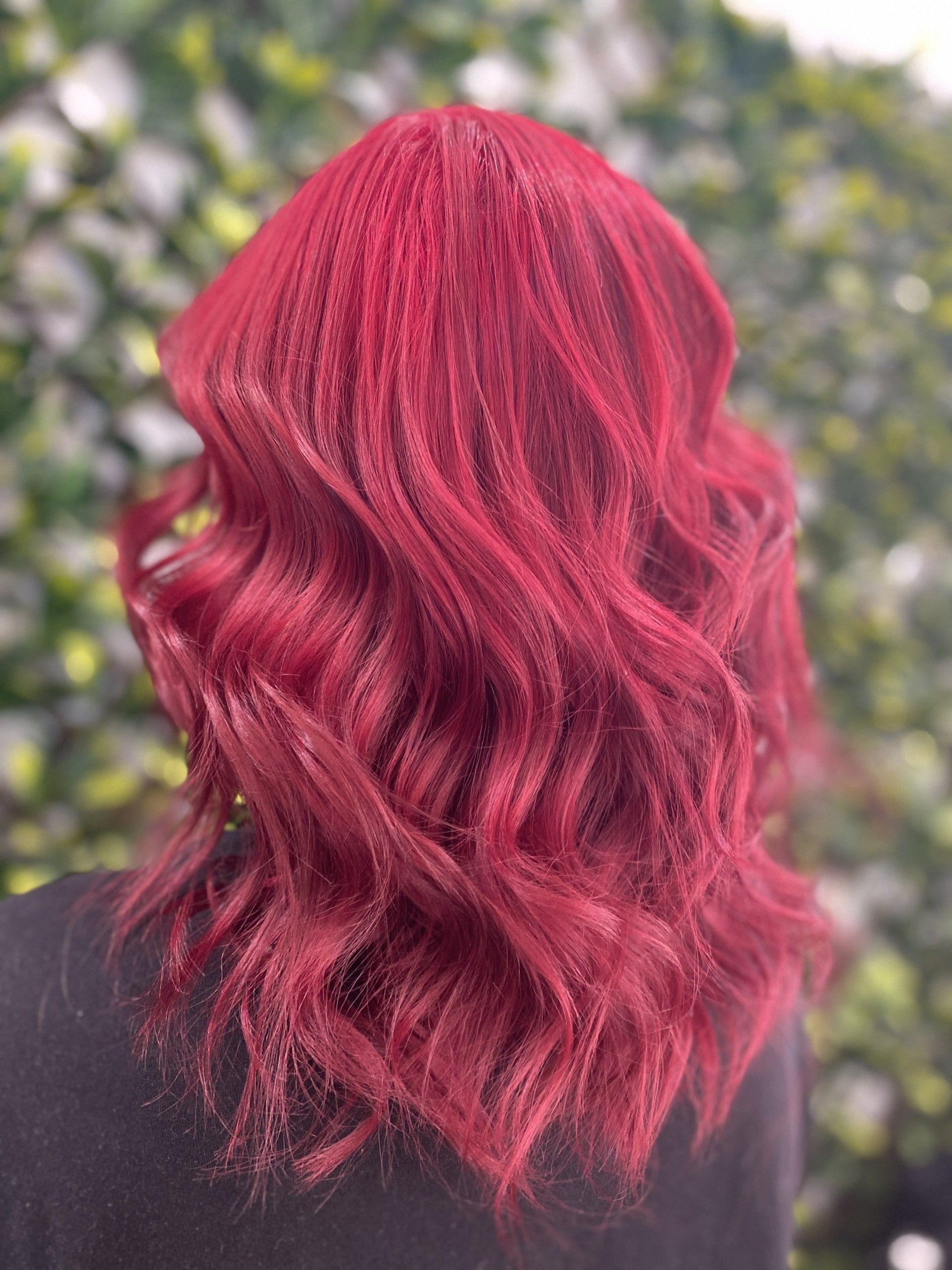 Woman with wavy, vibrant red hair, viewed from the back, against a green leafy background.