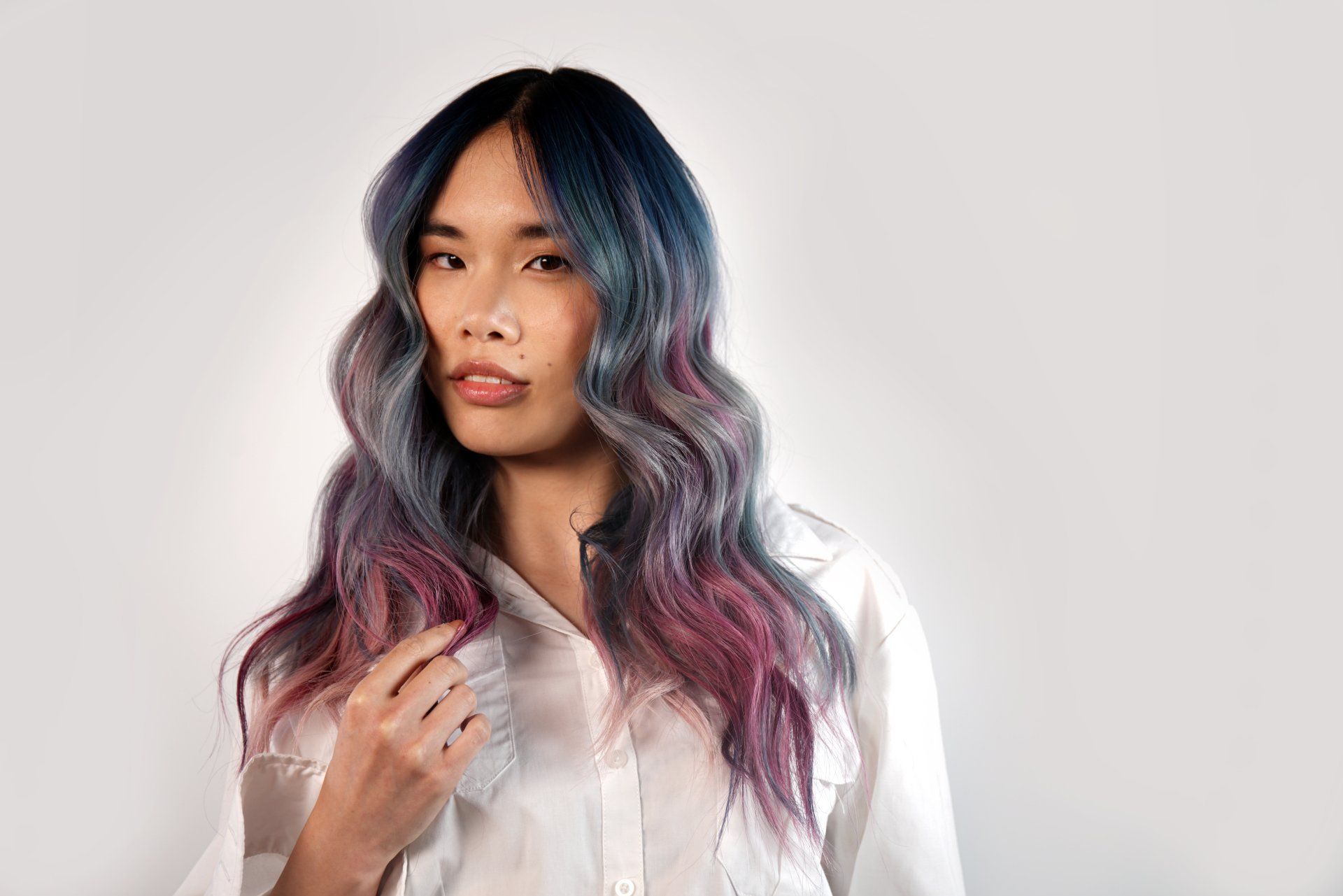 Woman with long, wavy hair in shades of blue, pink, and purple, wearing a white shirt, looking toward the viewer.