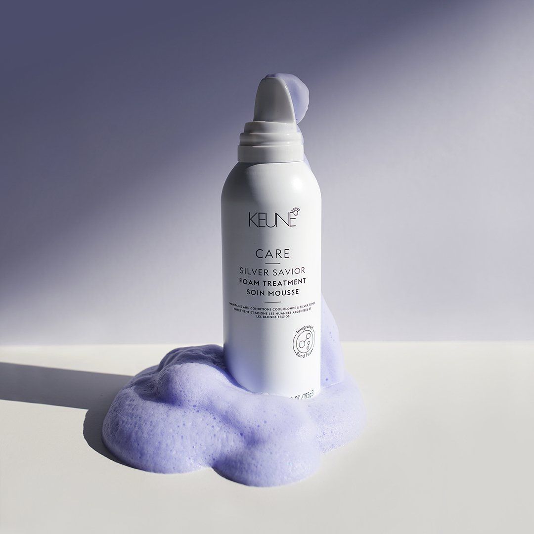 White bottle dispensing lavender-colored foam; product on a white surface, purple backdrop.