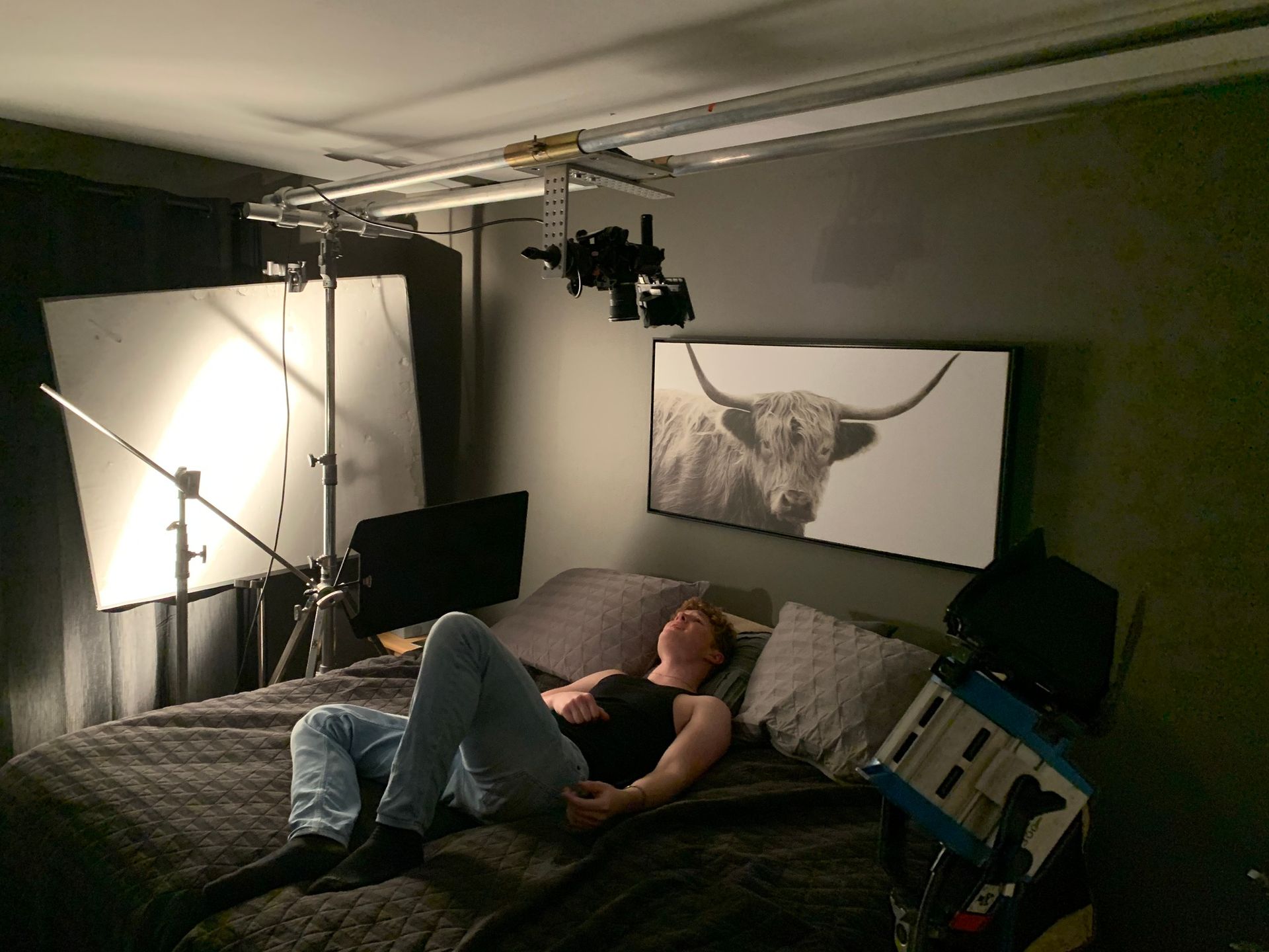 Person lying on a bed in a studio set, lit by bright overhead lights with a large bull portrait on the wall.