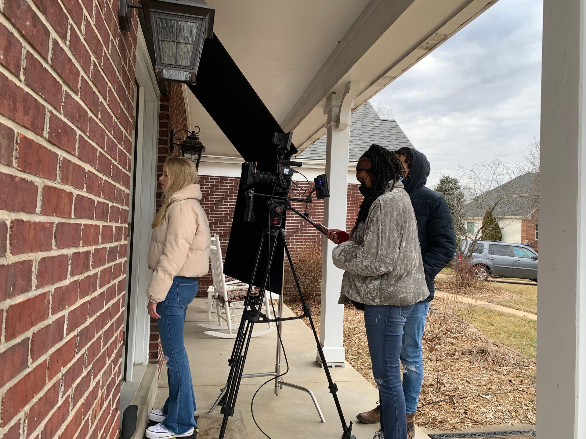 Three people filming by a brick house porch with a tripod and camera from the sidewalk