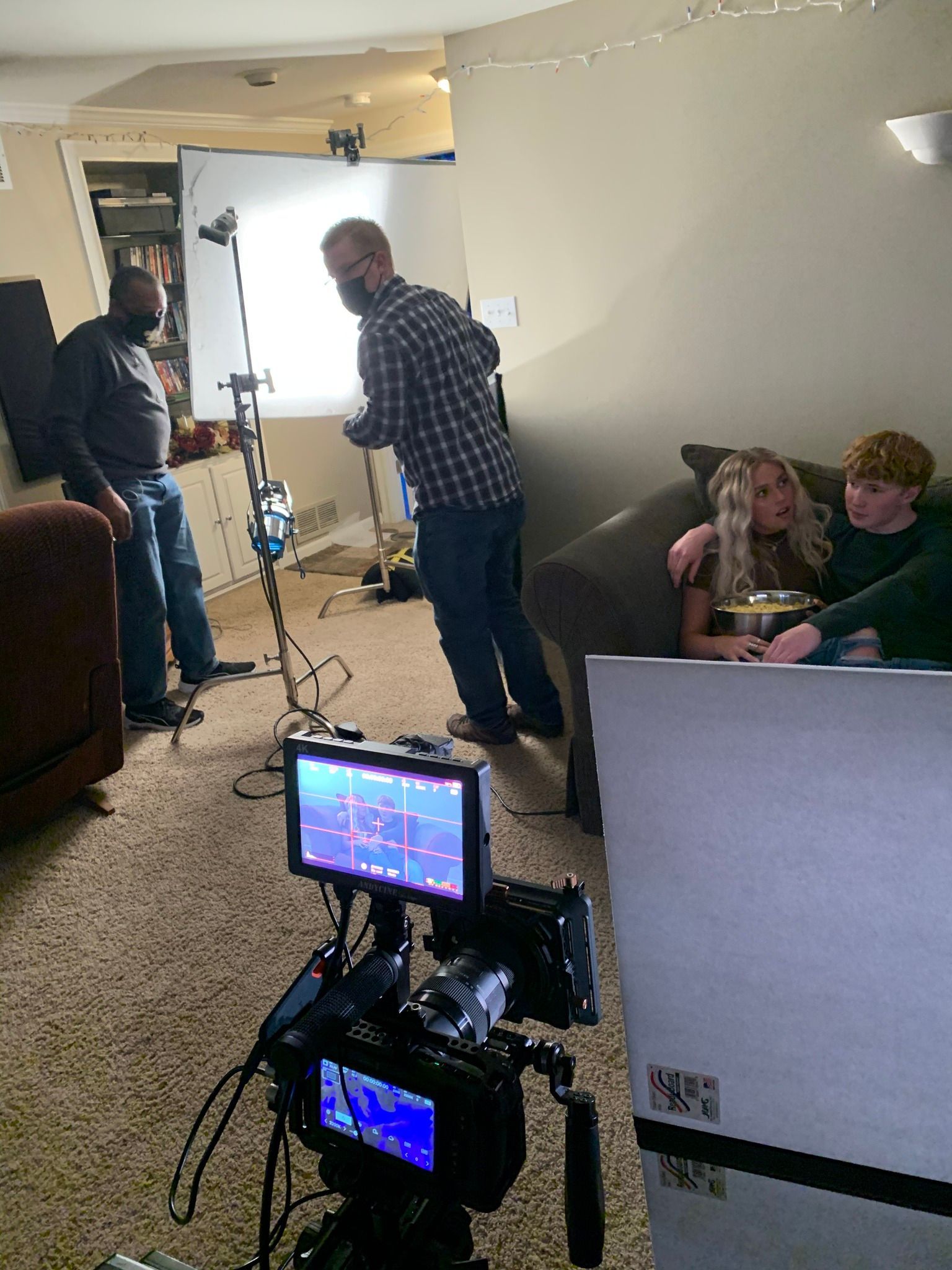 Film crew and lighting setup in a living room, with two people sitting on a couch in the background.