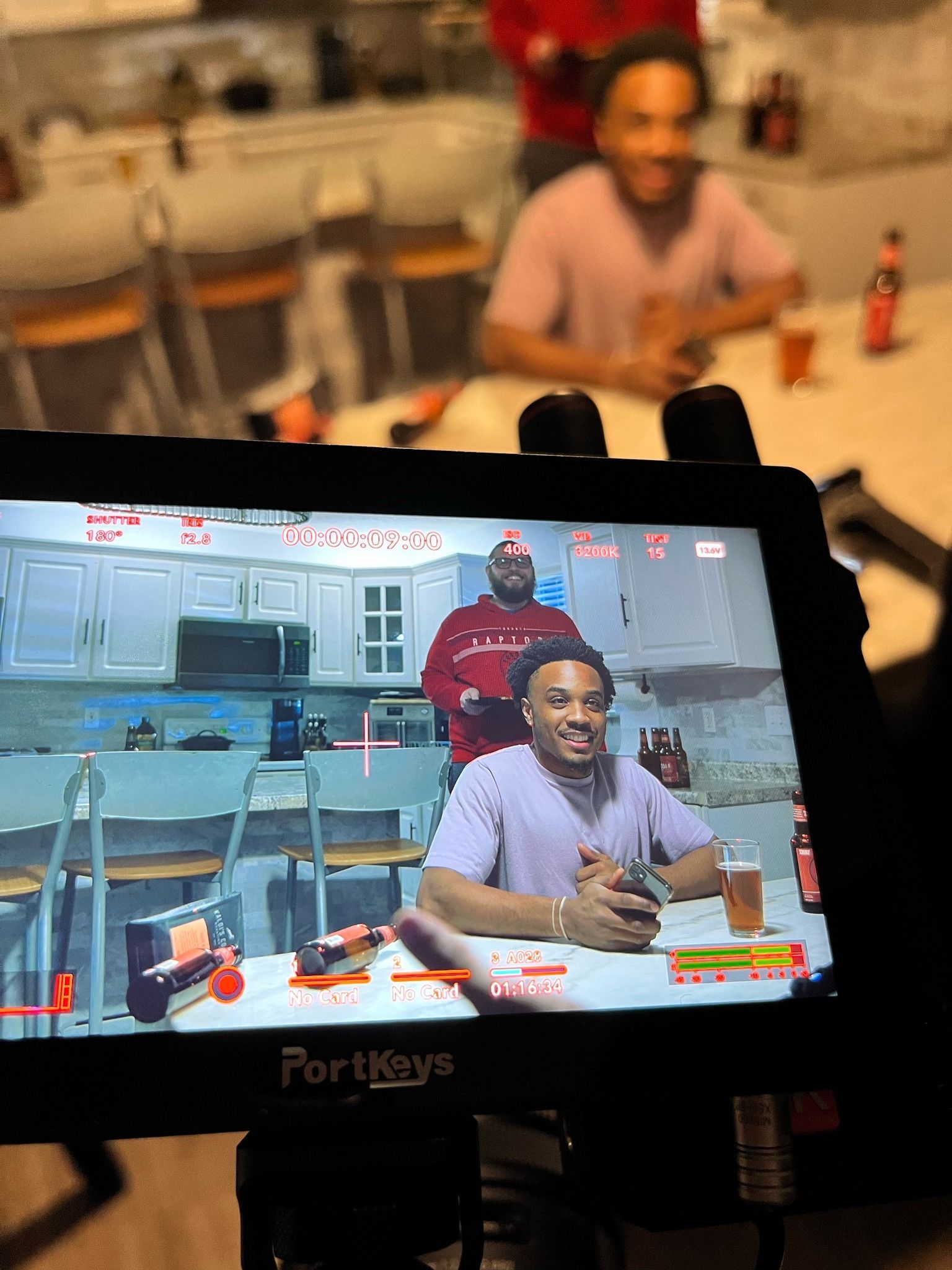 Camera screen shows a seated person smiling in a kitchen set, with a blurred real person in the background.