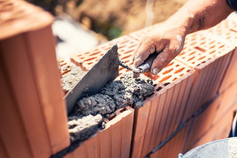 A Man Is Laying Bricks On A Wall With A Trowel — Heritage Bricklaying & Remedial Services In Toukley, NSW