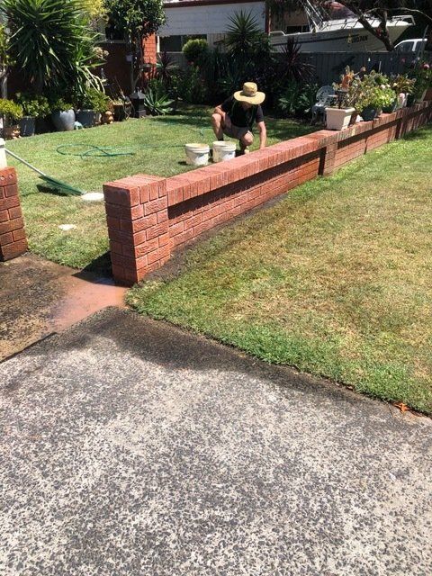 A Man In A Hat Is Kneeling On A Brick Wall In A Yard — Heritage Bricklaying & Remedial Services In Jilliby, NSW