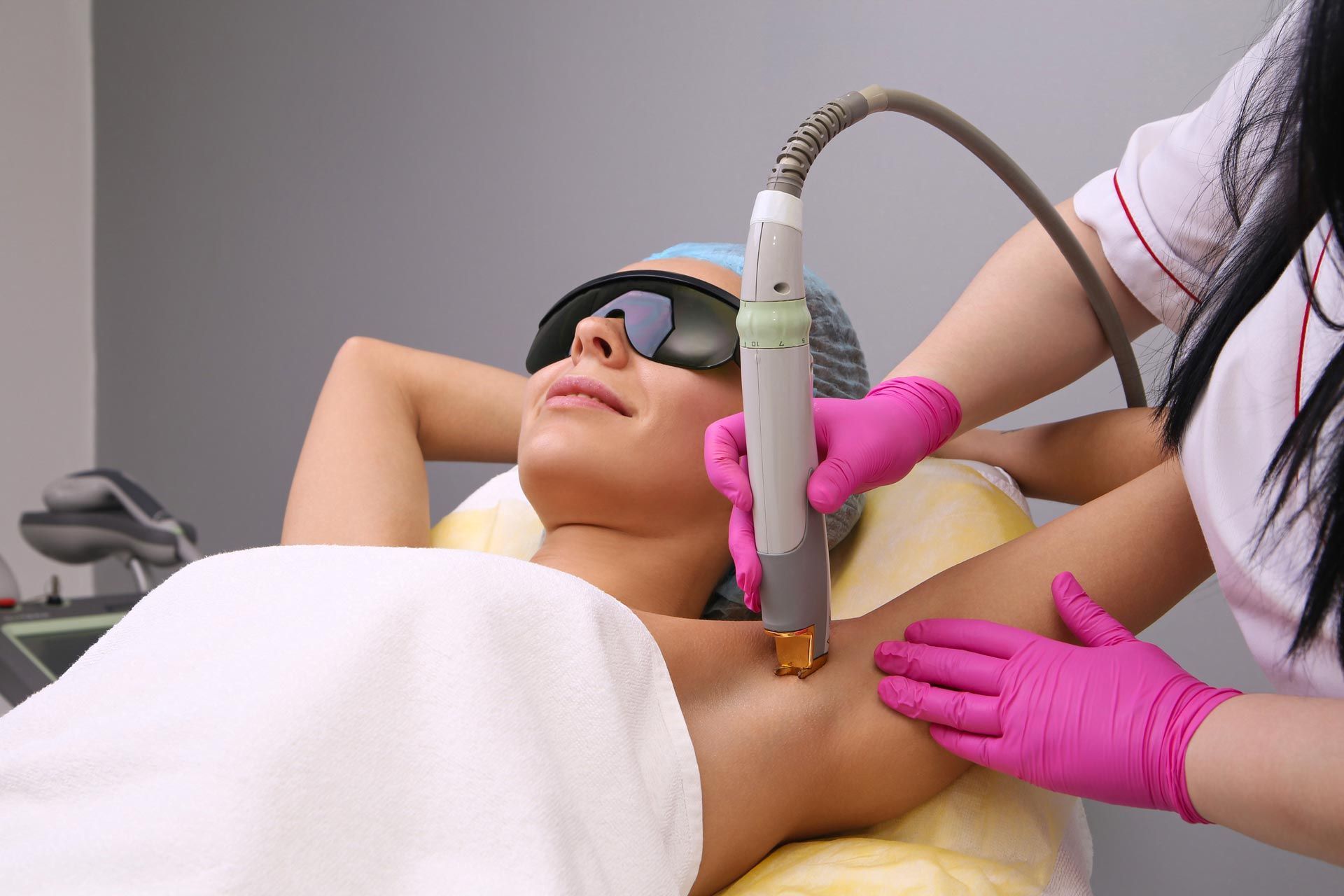 Esthetician performing laser treatment on a client’s underarm in a spa clinic, with protective goggles and pink gloves