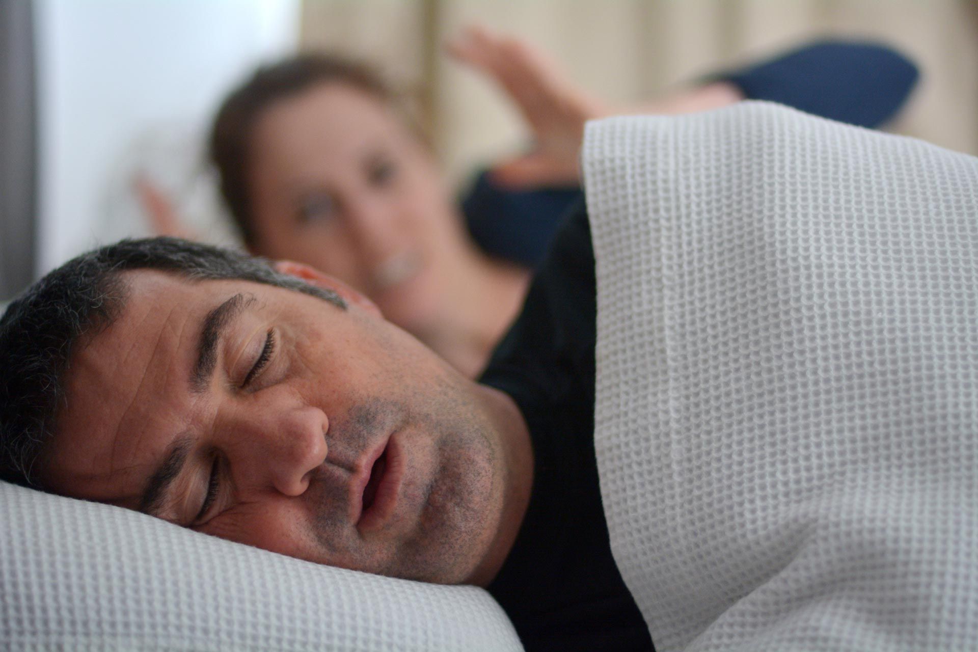 Man sleeping on a pillow under a white blanket, with a blurred person resting in the background