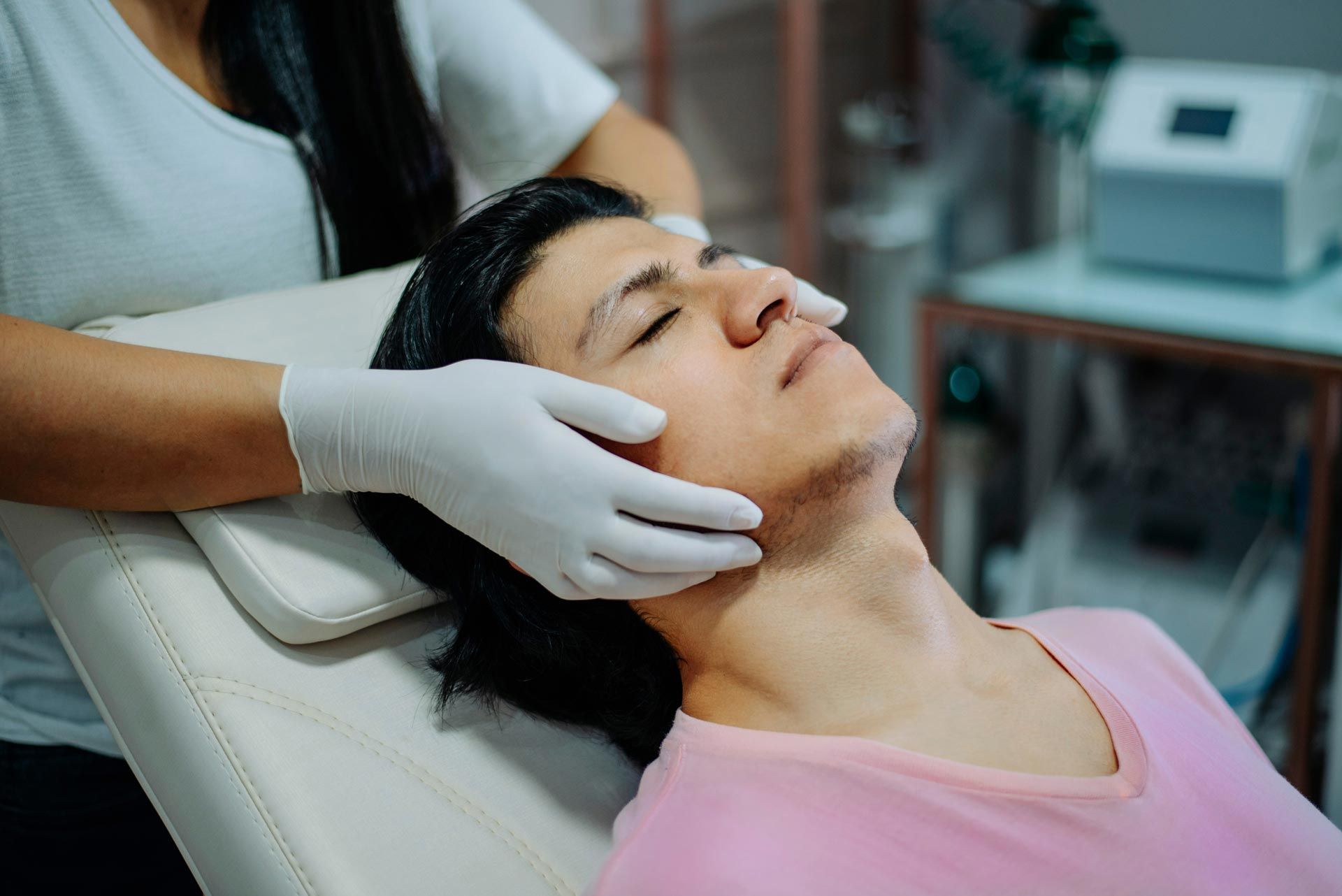 Gloved practitioner gently massages a reclining patient’s face in a clinic treatment room